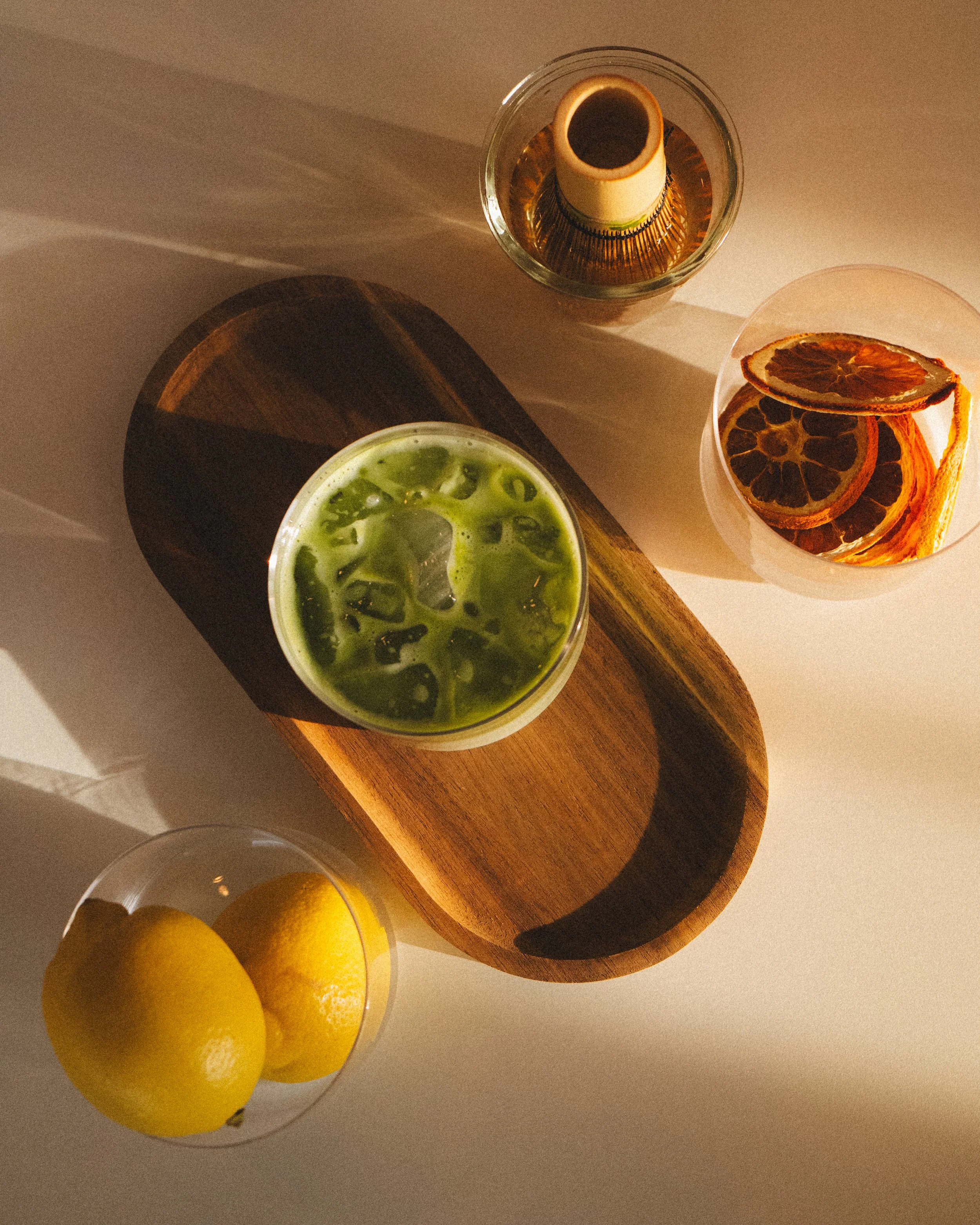 Top view of a glass with a drink and orange slices, a small bottle inside a glass bowl, and three lemons on a wooden board.