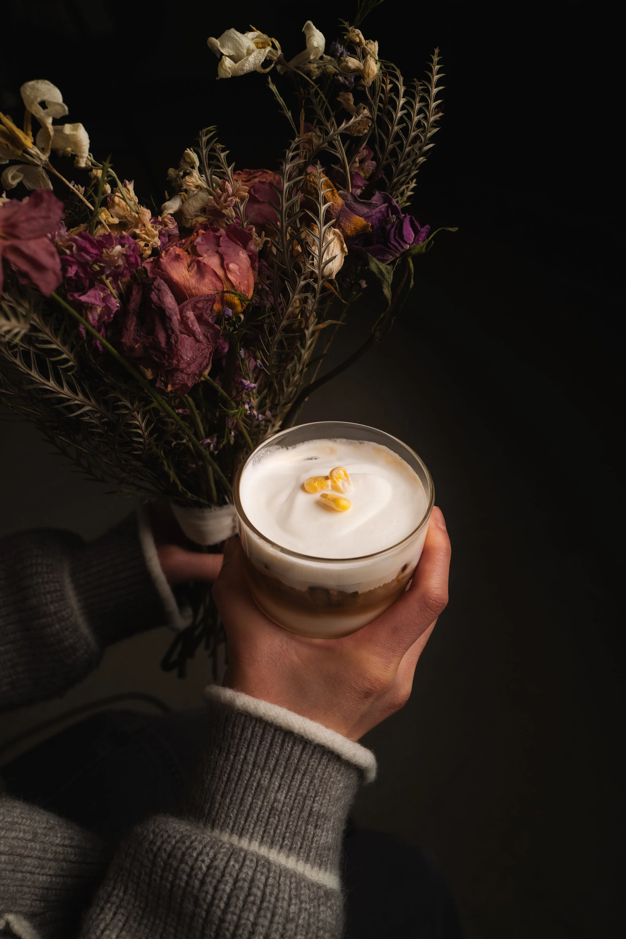 Close-up of a hand holding a layered dessert topped with whipped cream and yellow garnishes, with dried flowers and foliage in the background. Columbus, Ohio, David Kim Photography