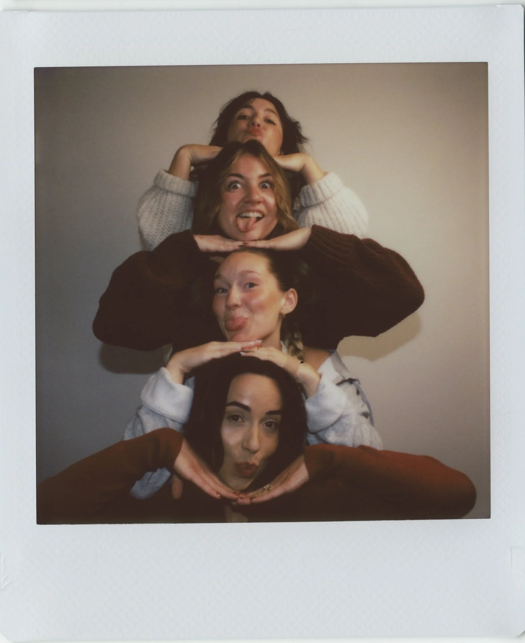 A group of five women sitting in a pile, making playful faces and hand gestures, with one woman at the top with curly hair and a white sweater, and others forming hearts and peace signs with their hands.