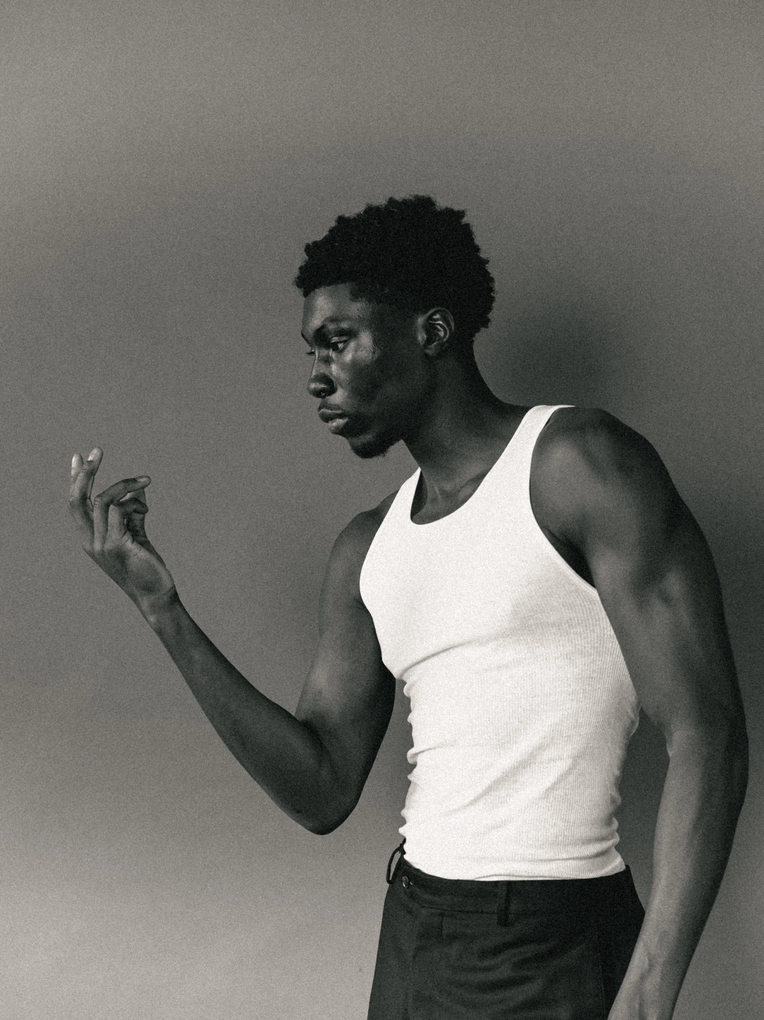 A black-and-white image of a man with dark skin and textured hair, wearing a white tank top and black pants, looking downwards with his left hand raised near his face against a plain background.