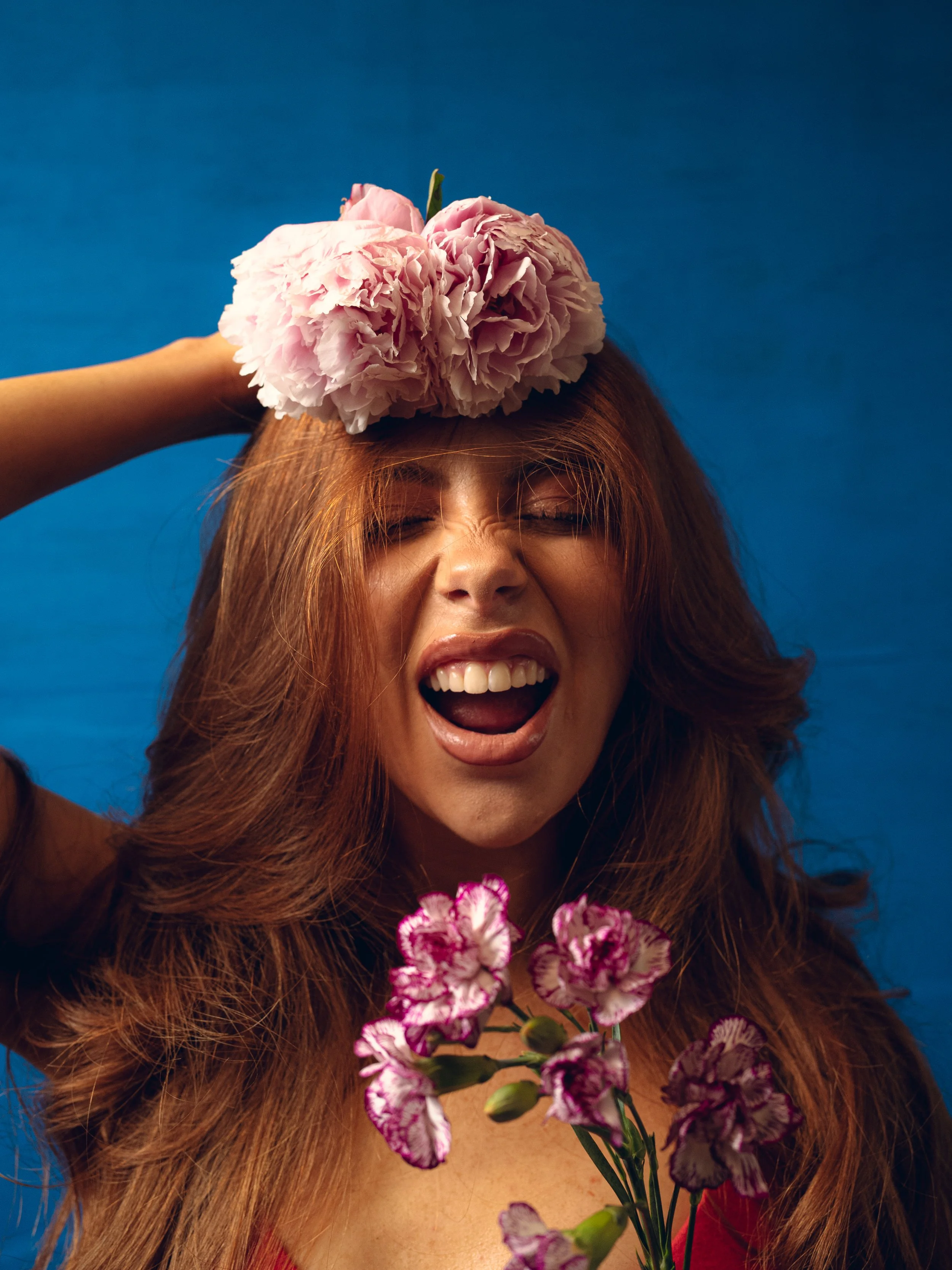 Woman with long red hair holding pink and purple flowers, smiling with eyes closed, against a blue background. Columbus, Ohio, David Kim Photography