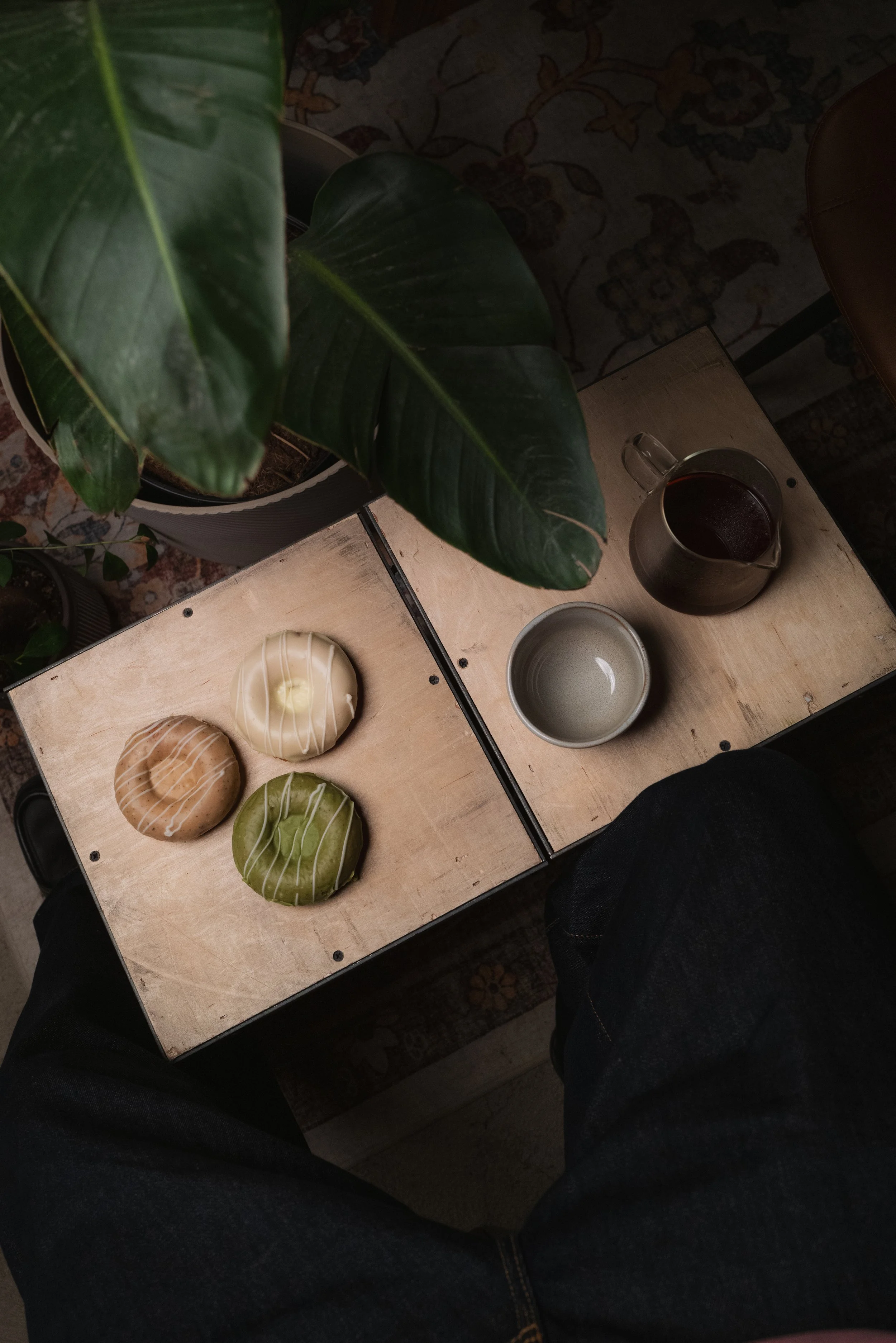 A person sitting at a wooden table with several decorated donuts, a small potted plant, a cup of coffee, and a glass mug of dark beverage. Columbus, Ohio, David Kim Photography