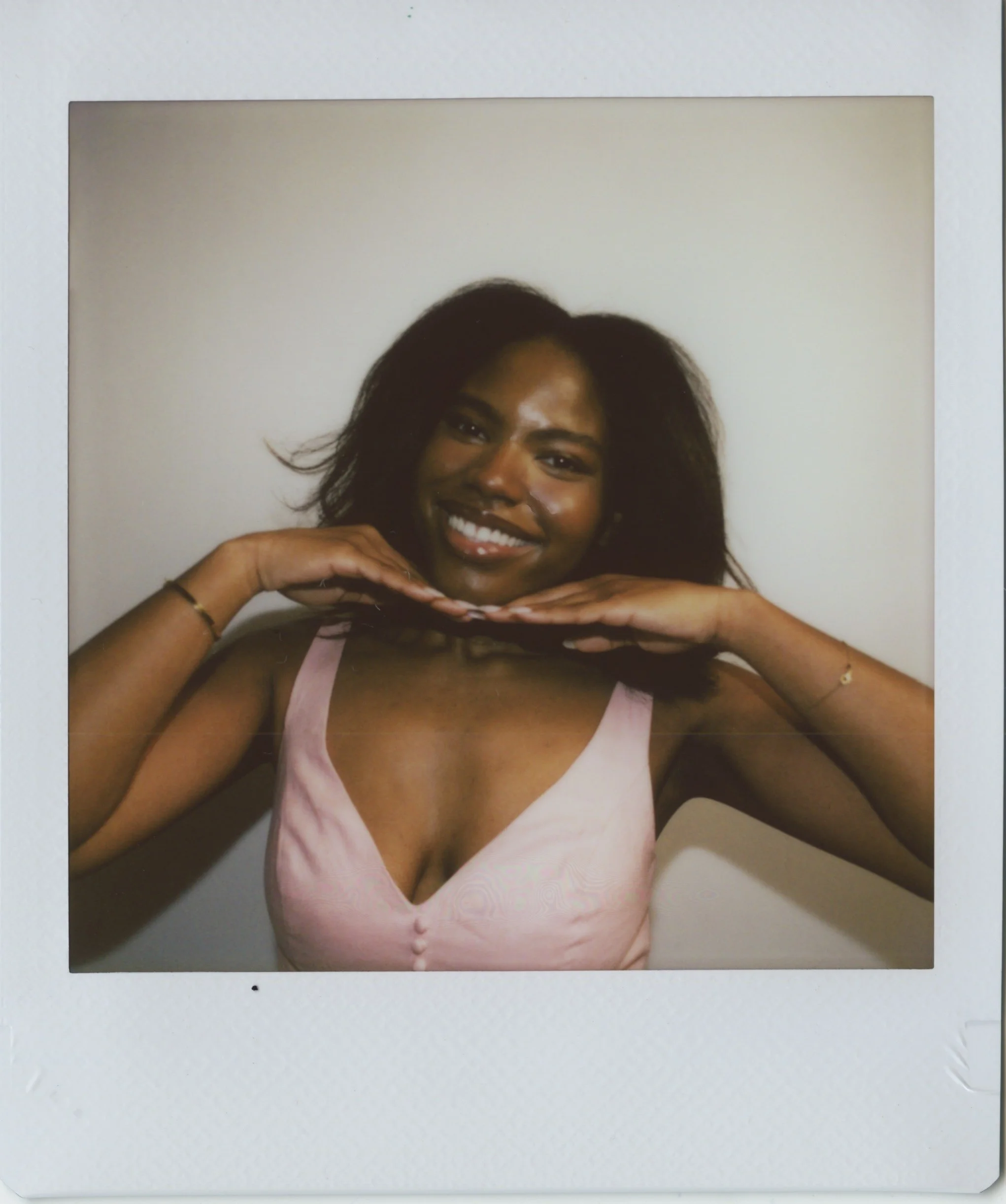Photograph of a woman with dark hair, smiling, resting her chin on her hands, wearing a pink top, against a light-colored background.