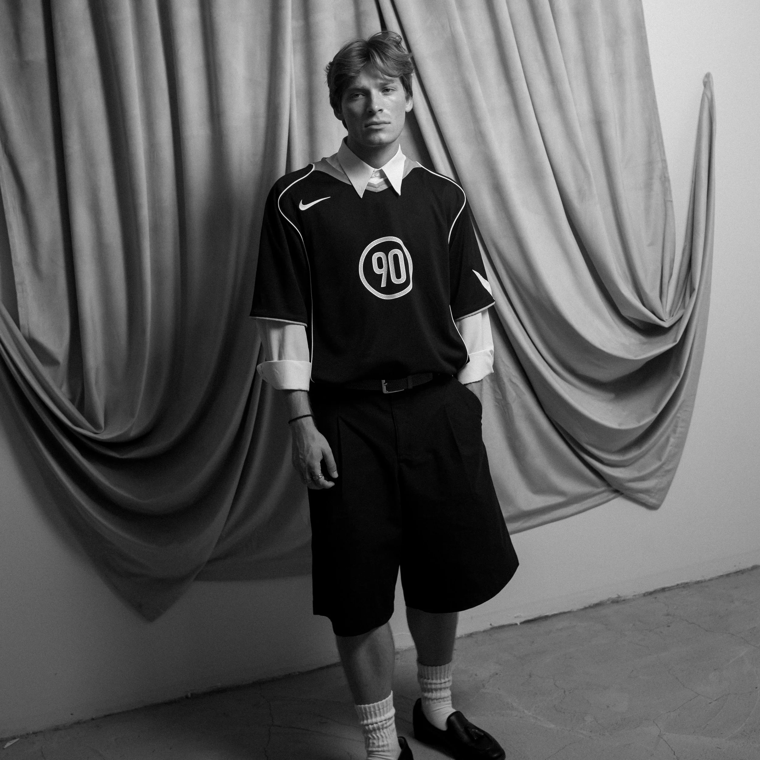Black and white photo of a young man standing in front of a curtain, wearing a vintage football jersey with the number 90, shorts, knee-high socks, and loafers. Columbus, Ohio, David Kim Photography