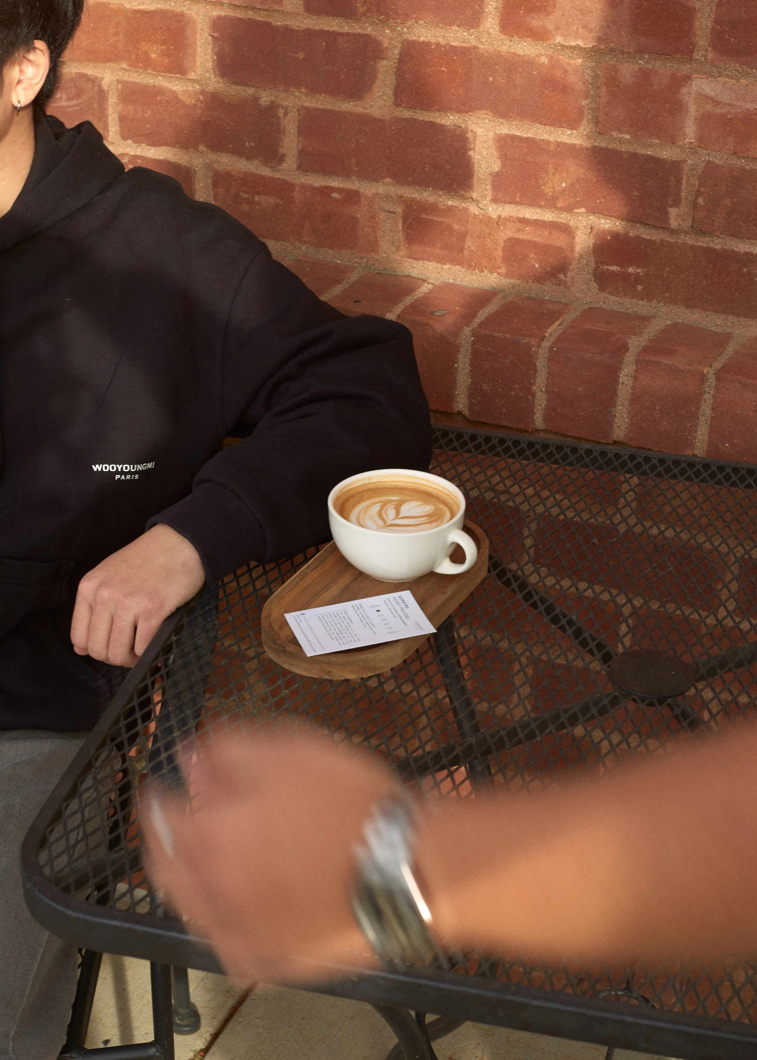 Person sitting at a black metal outdoor table with a cup of coffee and a receipt, against a brick wall background. Columbus, Ohio, David Kim Photography