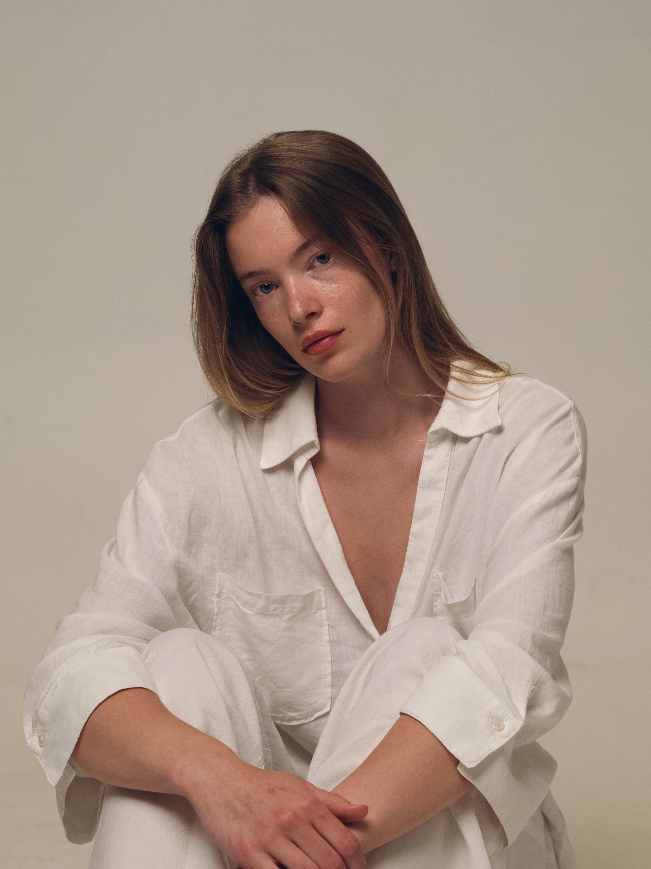 A woman with red hair, blue eyes, and freckles, wearing a white shirt and sitting against a plain background.