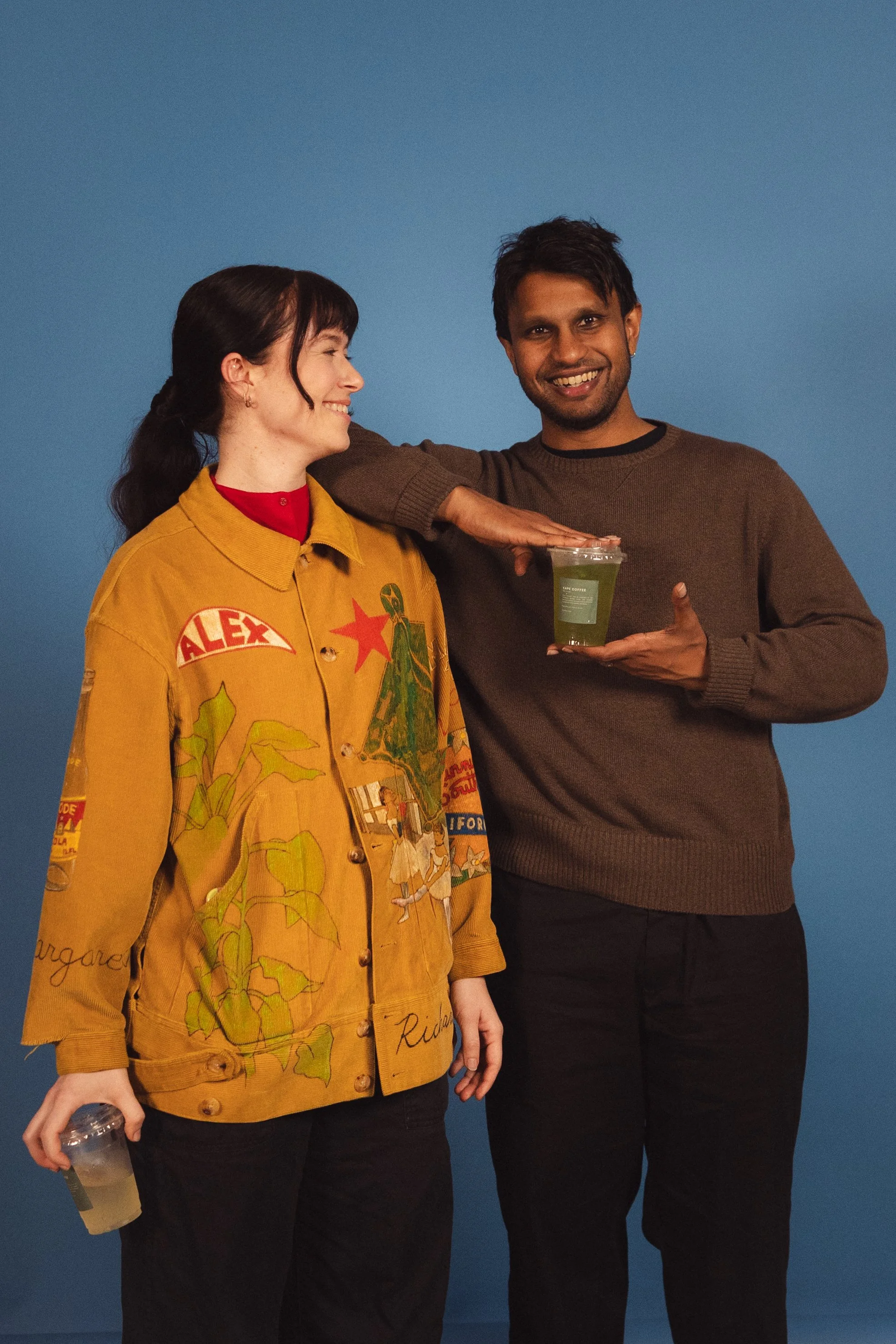 A woman with black hair tied back, wearing a mustard yellow jacket with colorful patches and black pants, is smiling at a man. The man, with short dark hair and earrings, is smiling back and holding a clear plastic cup with green drink, standing agai
