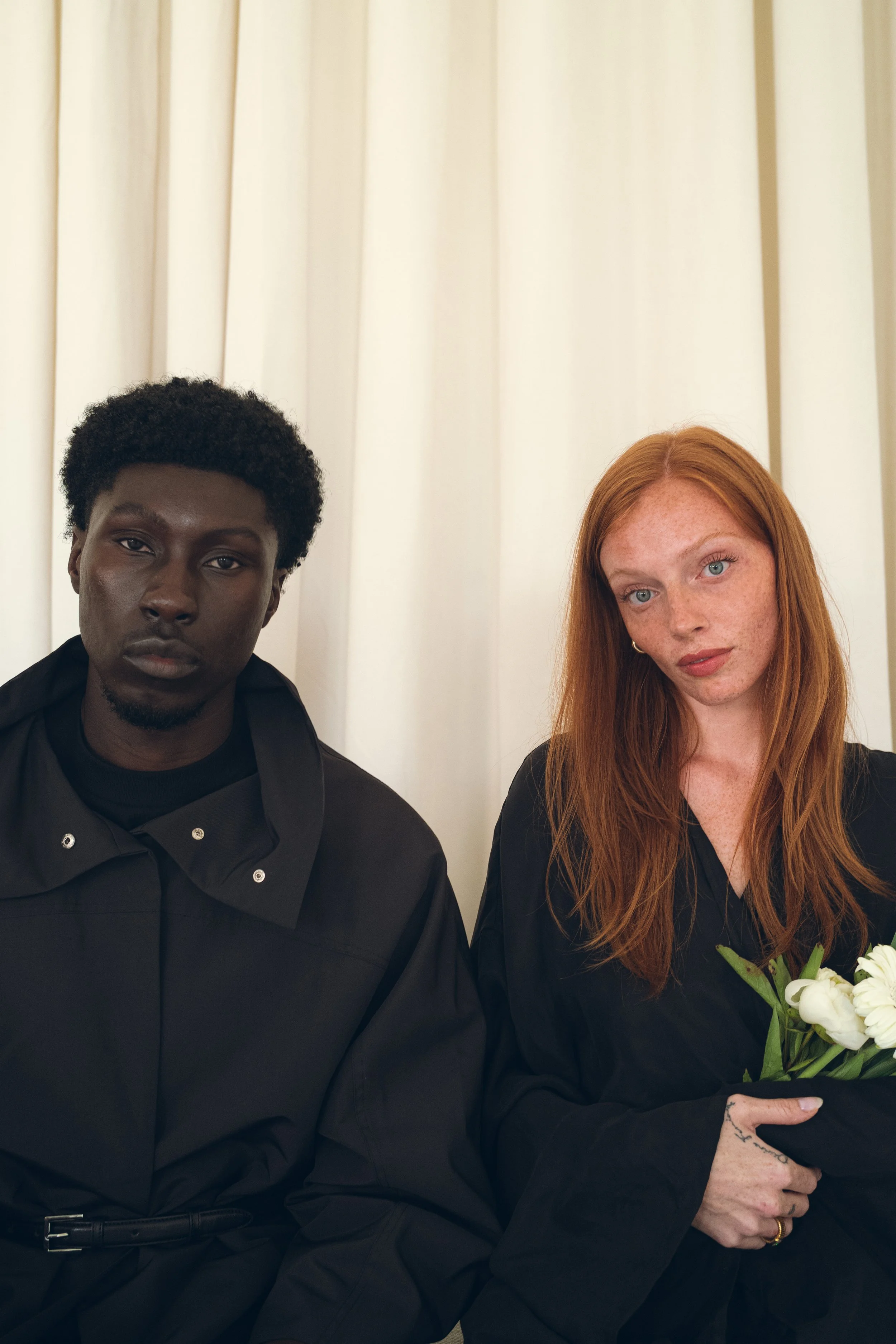 A young Black man and a young red-haired woman sit side by side against a cream-colored curtain. The man has short curly hair and wears a black shirt with a buttoned collar. Columbus, Ohio, David Kim Photography