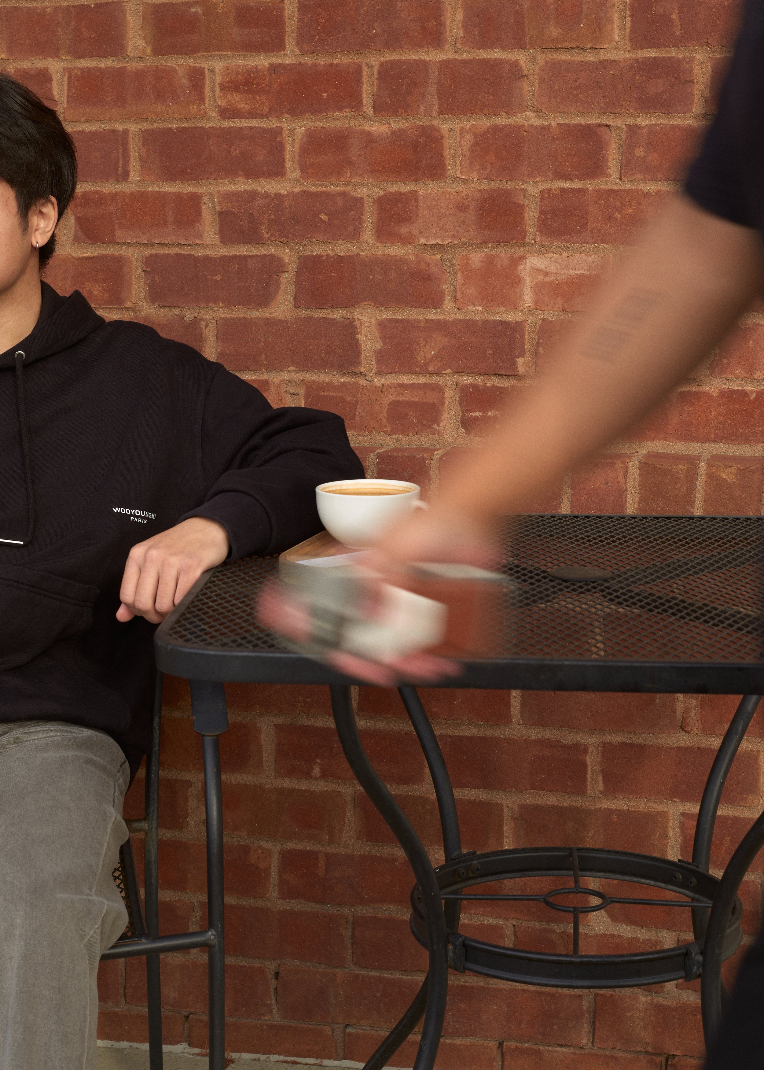 Part of a person's face, arm, and lap are visible, sitting at a black metal outdoor table with a white coffee cup and saucer, against a red brick wall background. Columbus, Ohio, David Kim Photography