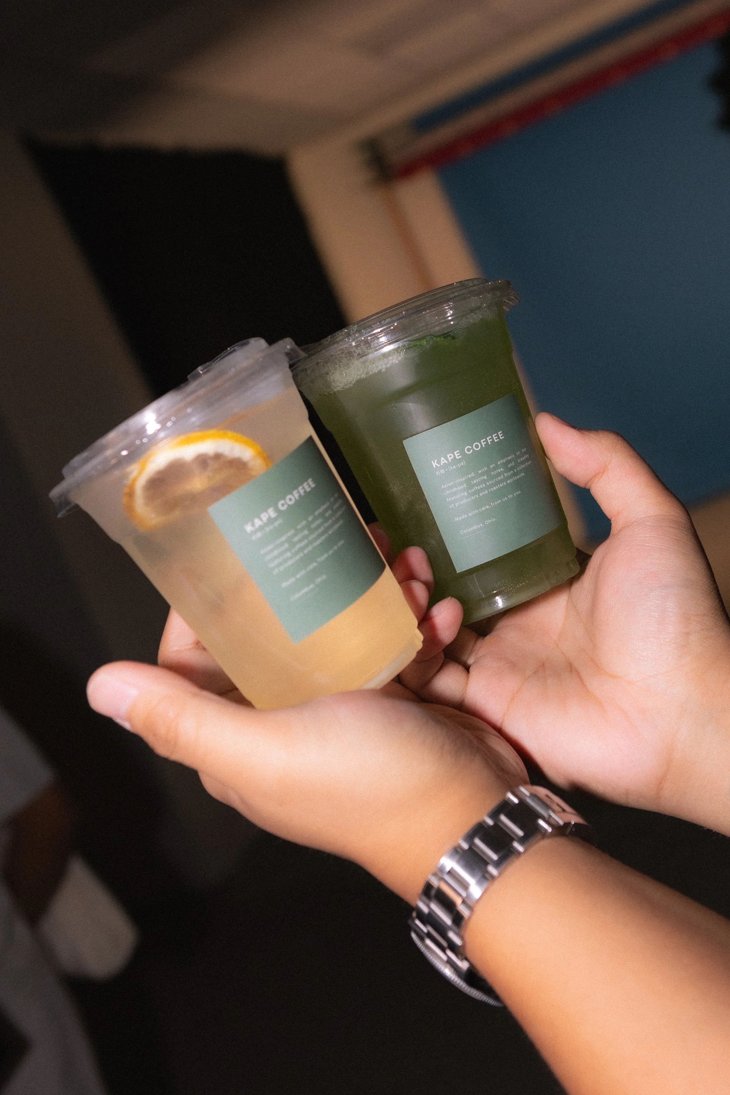 A person's hand holding two plastic cups filled with cold beverages, one with a lemon slice and the other with a green drink, labeled 'KAPE COFFEE', in an indoor setting.