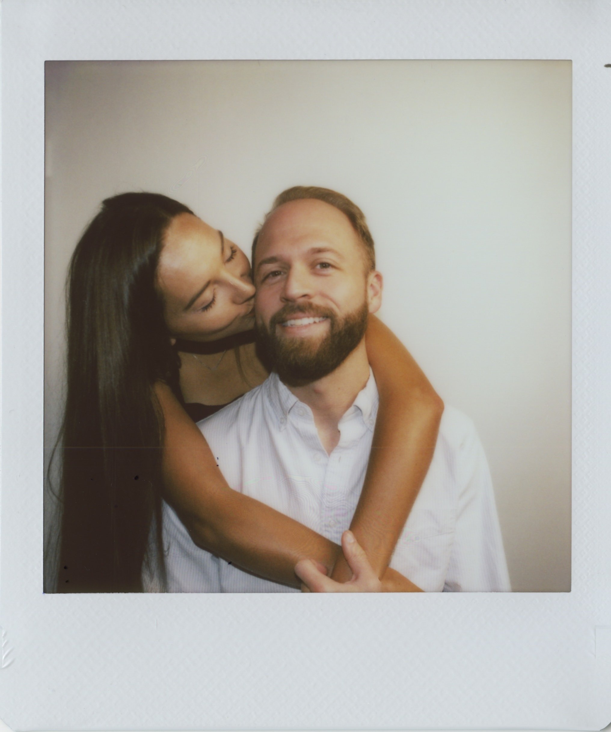 A woman with long dark hair kissing a smiling man with a beard and short blond hair, who is wearing a white shirt, against a plain background.