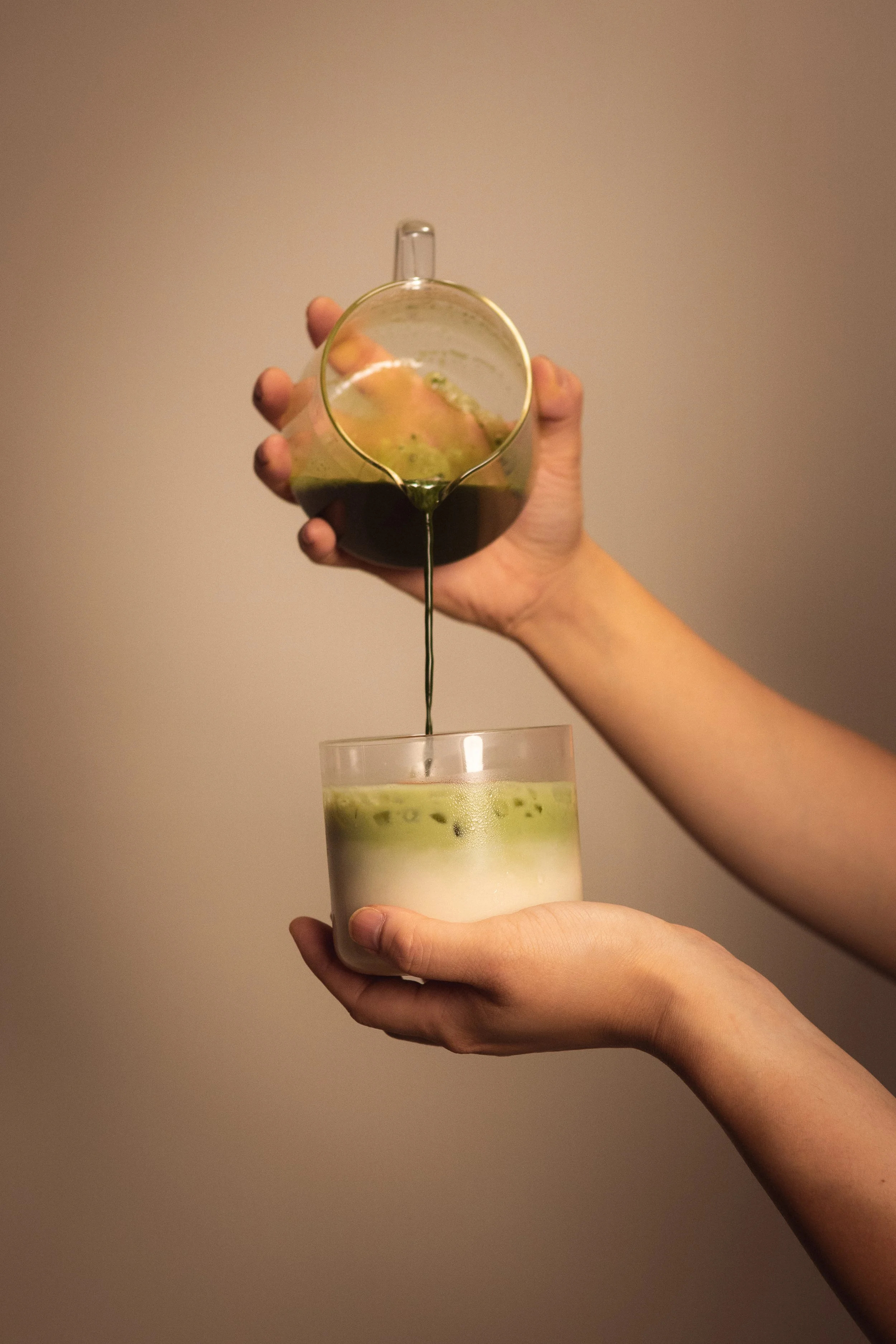 A person pours a dark green matcha latte from a small glass pitcher into a glass of layered matcha latte with milk on the bottom and matcha in the middle. Columbus, Ohio, David Kim Photography