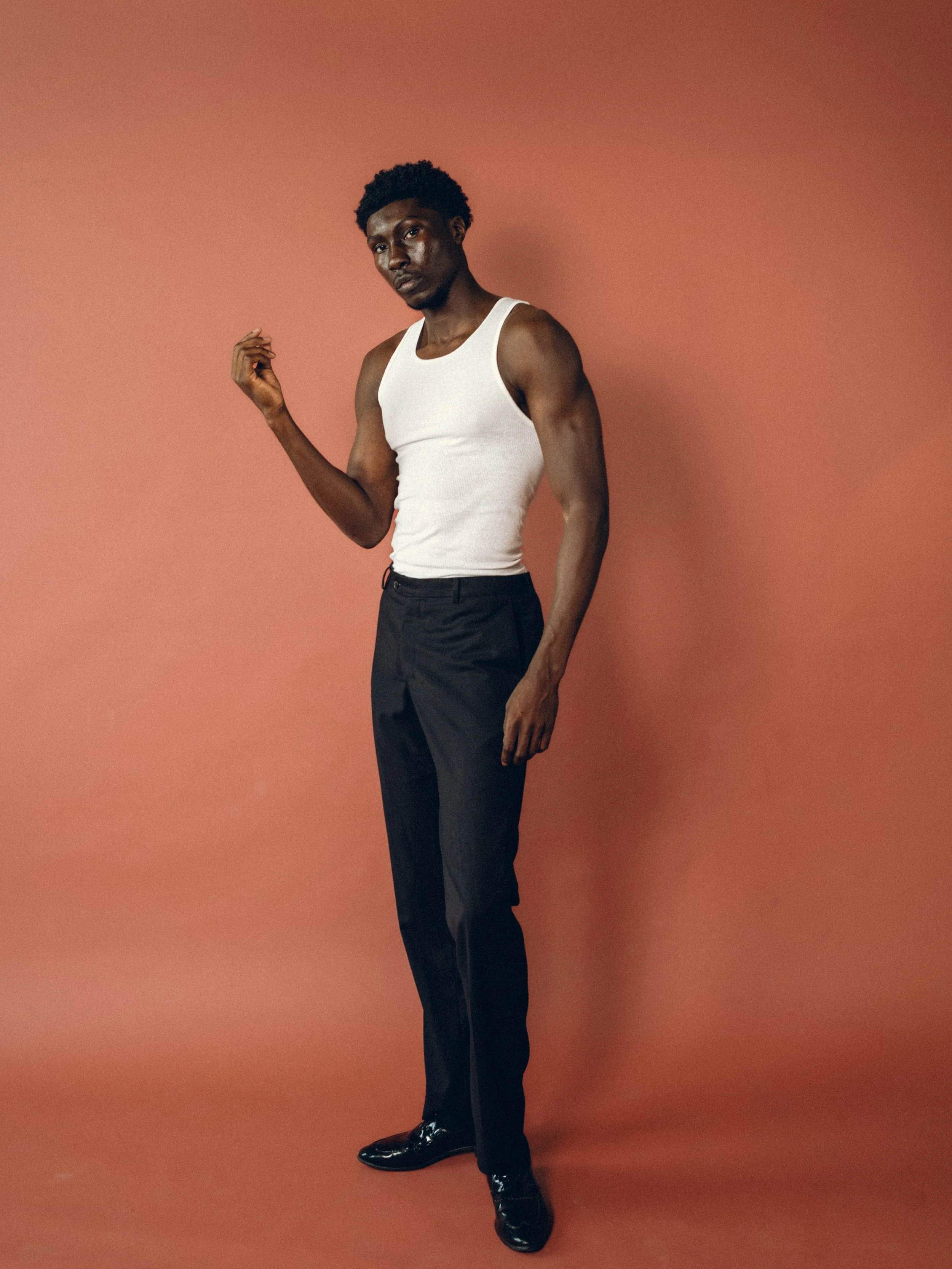 A young man with dark skin and curly hair stands against a reddish-brown background. He is wearing a white sleeveless shirt, black pants, and shiny black shoes. He is looking directly at the camera with a serious expression, with one arm bent holding