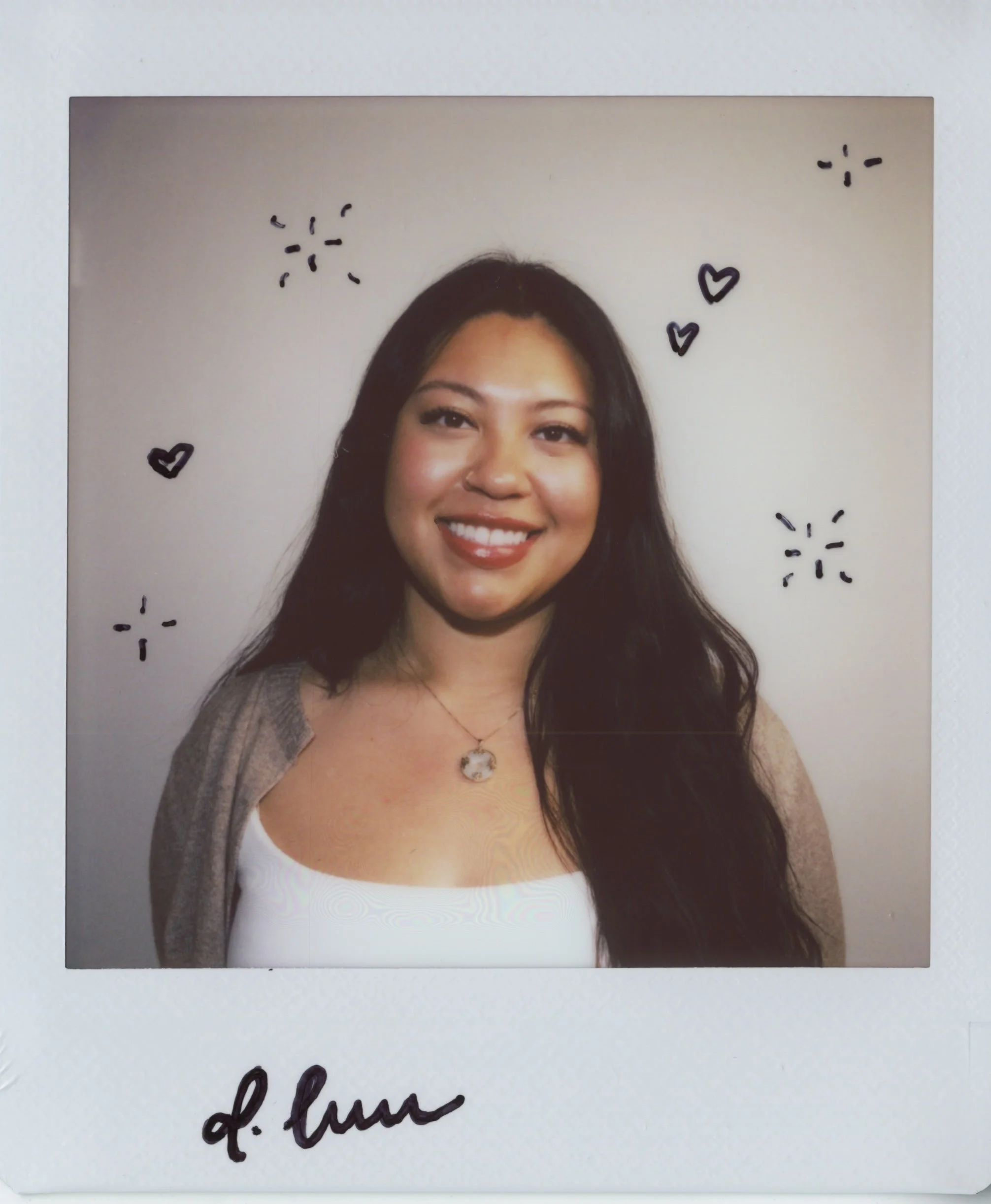 A smiling woman with long dark hair, wearing a necklace with a round pendant. The photo is decorated with small black drawings of hearts and sparkles, and has handwritten text underneath.
