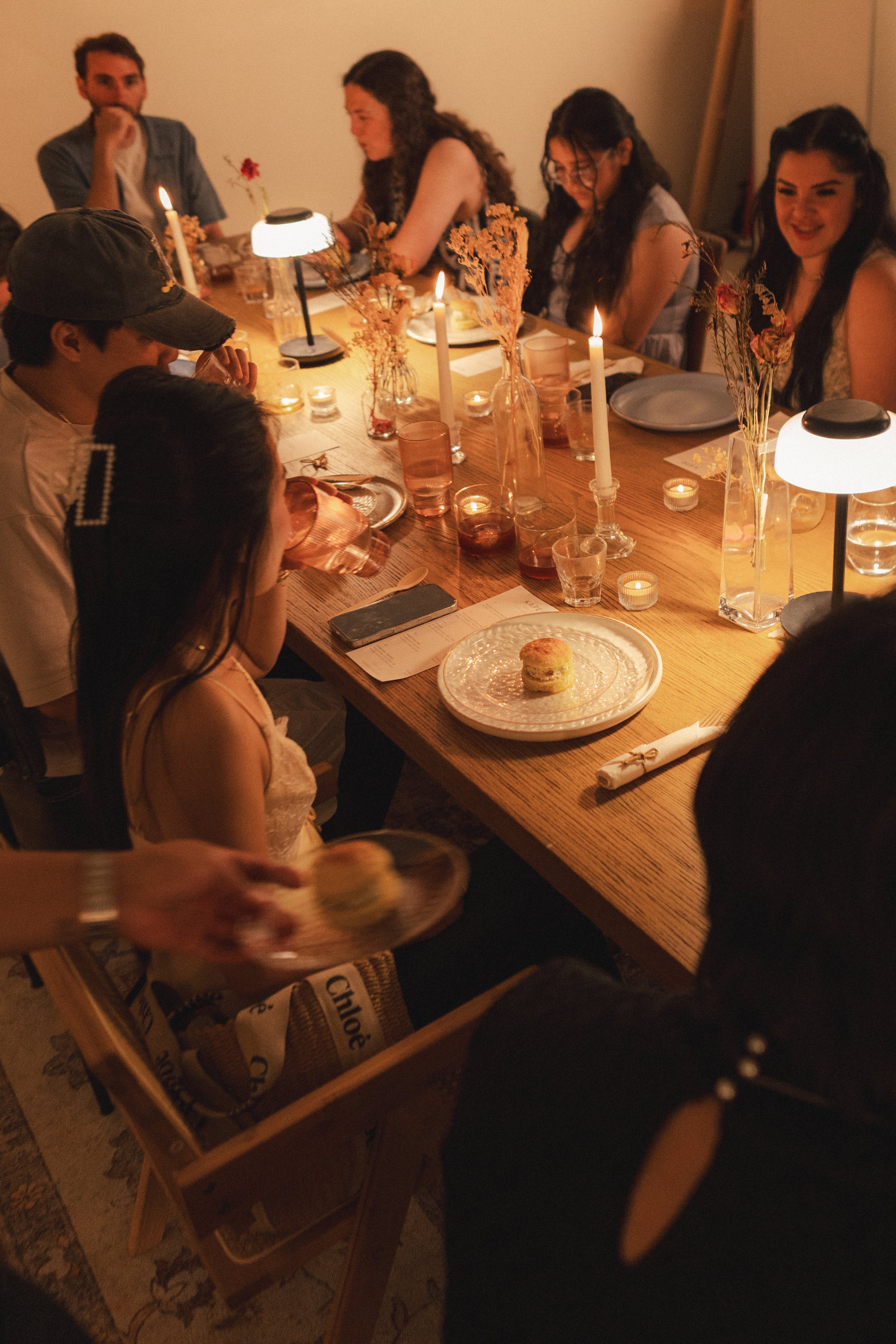 A group of people sitting around a wooden dining table decorated with candles, dried flowers, and plates, enjoying a meal in a cozy, dimly lit setting.