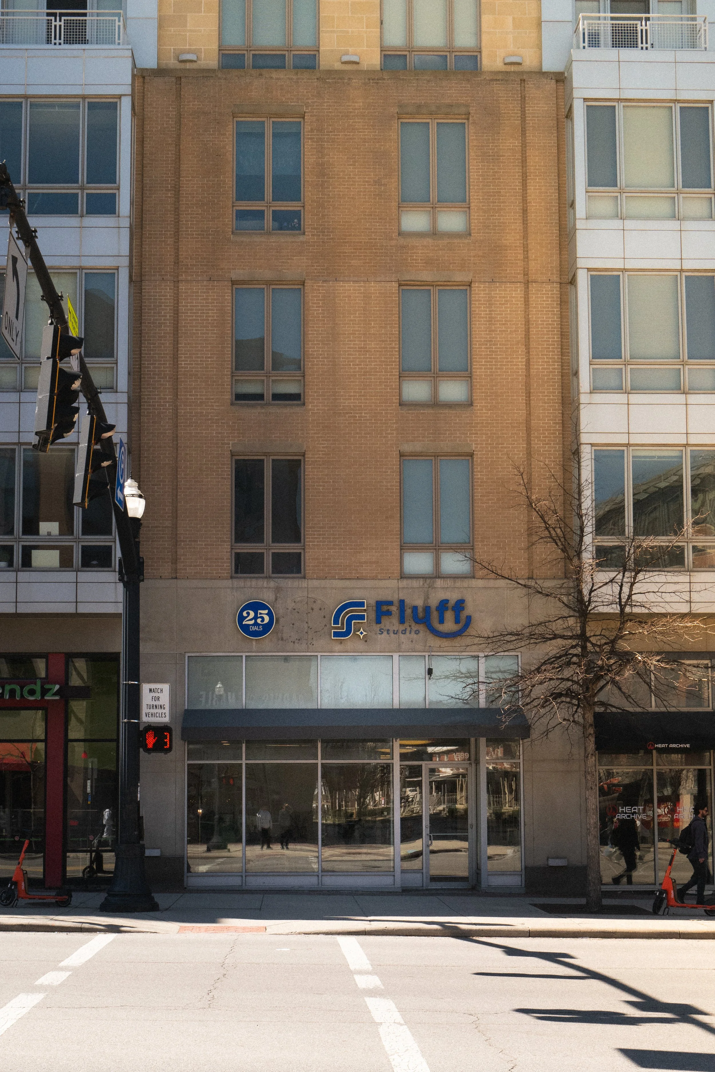 An urban scene with a brick and glass building and a storefront with glass doors across the street. There are parking spots, a streetlight, and a traffic signal. Pedestrians are visible through the glass doors and on the sidewalk. Columbus, Ohio, Dav