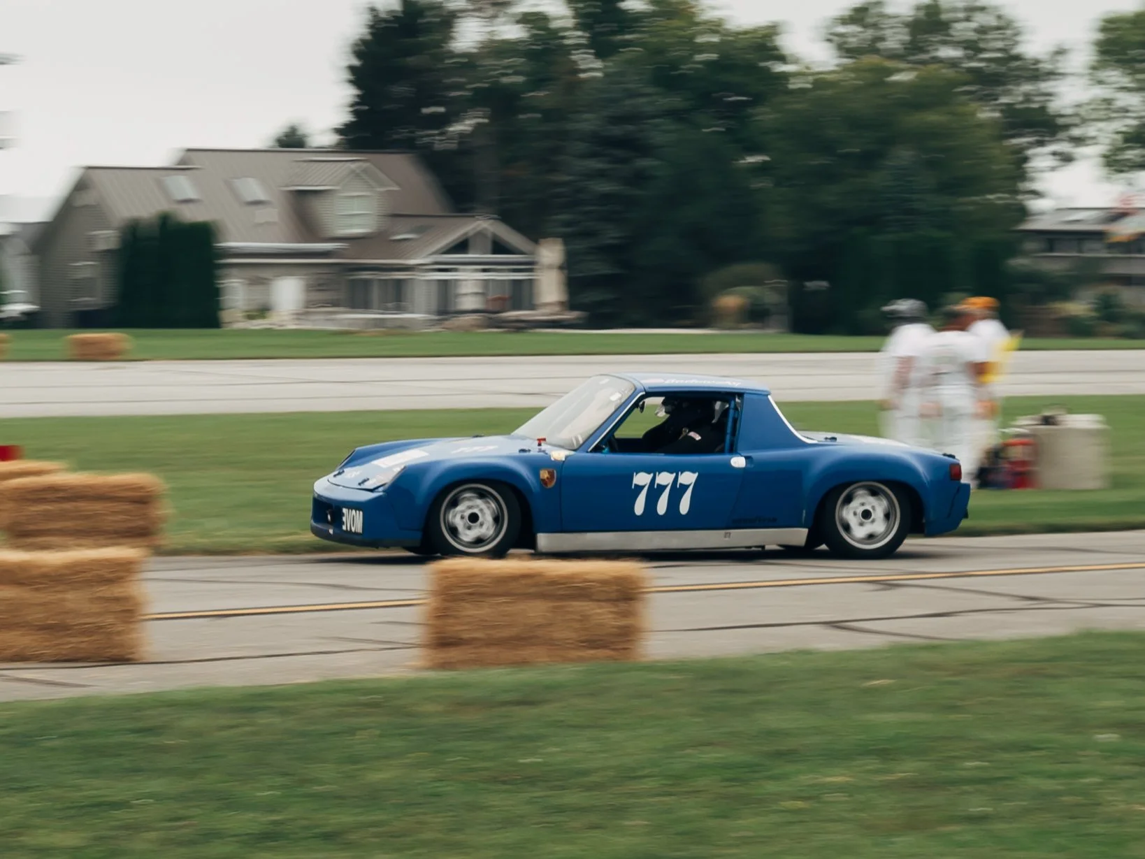A blue race car with the number 777 on its side driving on a race track lined with hay bales. There are a few people standing on the grass beside the track near some equipment and a house in the background. The scene appears to be at a racing event, 