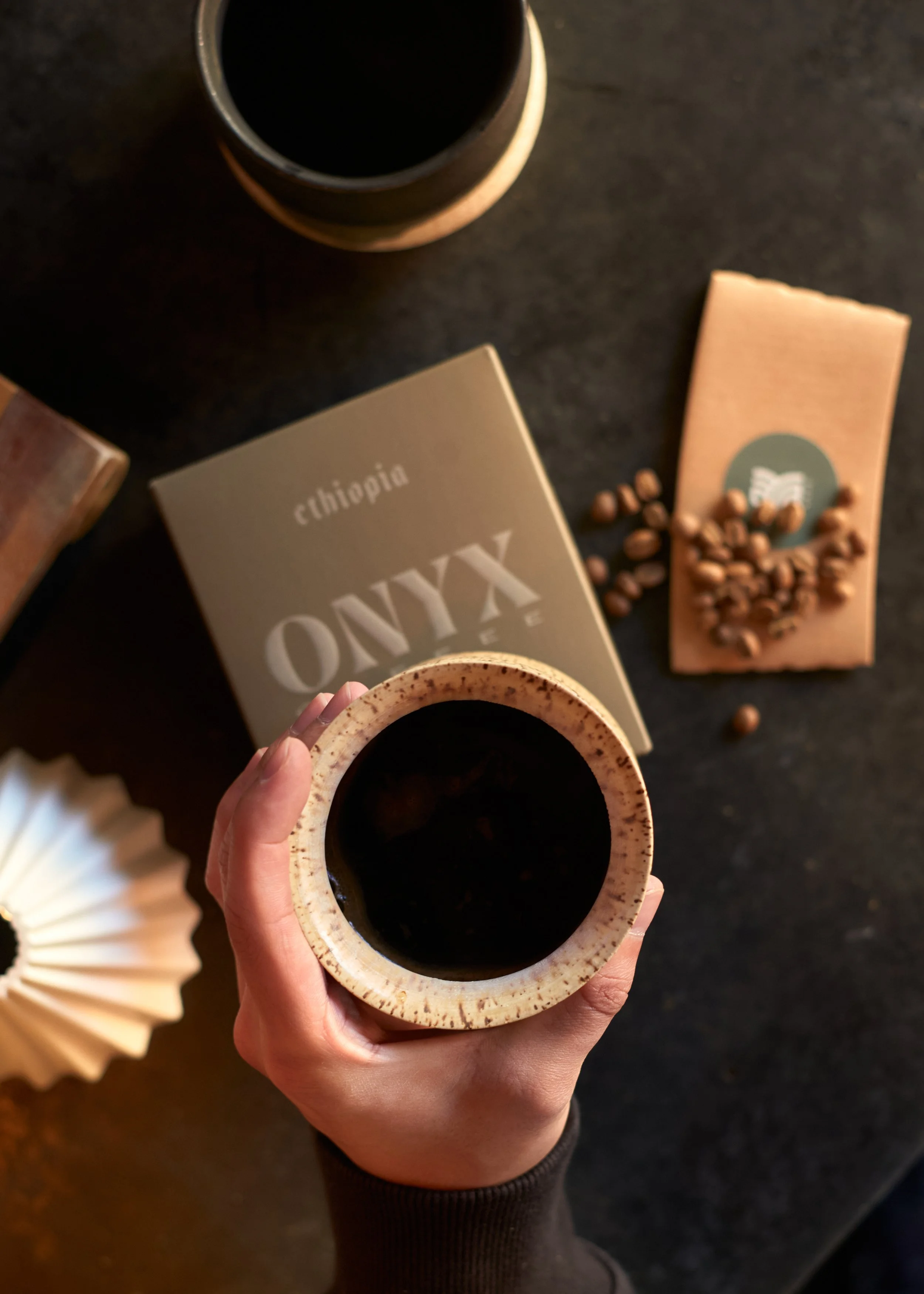 A hand holding a cup of black coffee over a dark surface, with a box of coffee beans and a pink coffee bar on the right side, and a book titled 'Culinary' partially visible in the background.