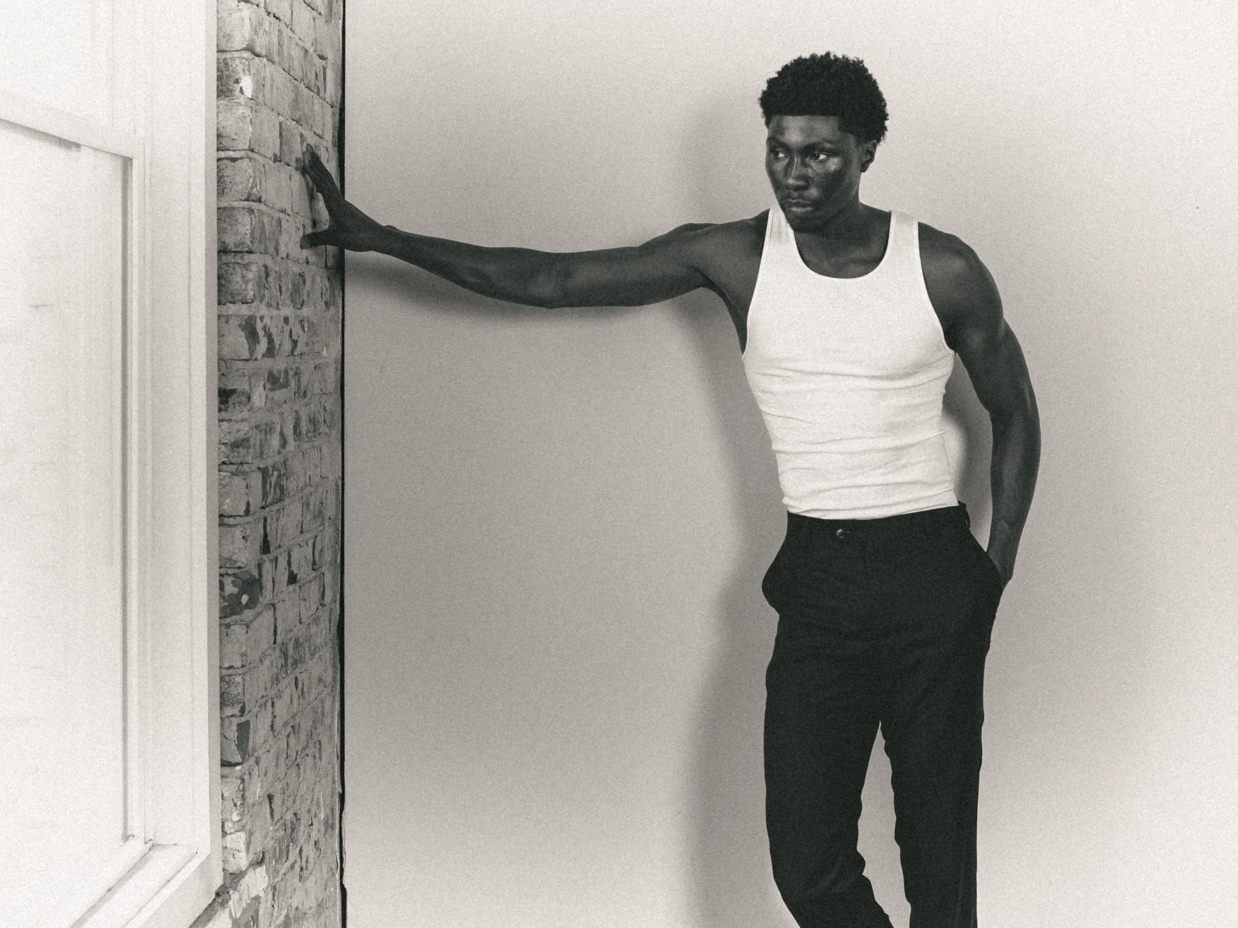 A young Black man with curly hair leaning against a wall with a brick section, wearing a white tank top and black pants, looking thoughtfully to the side.