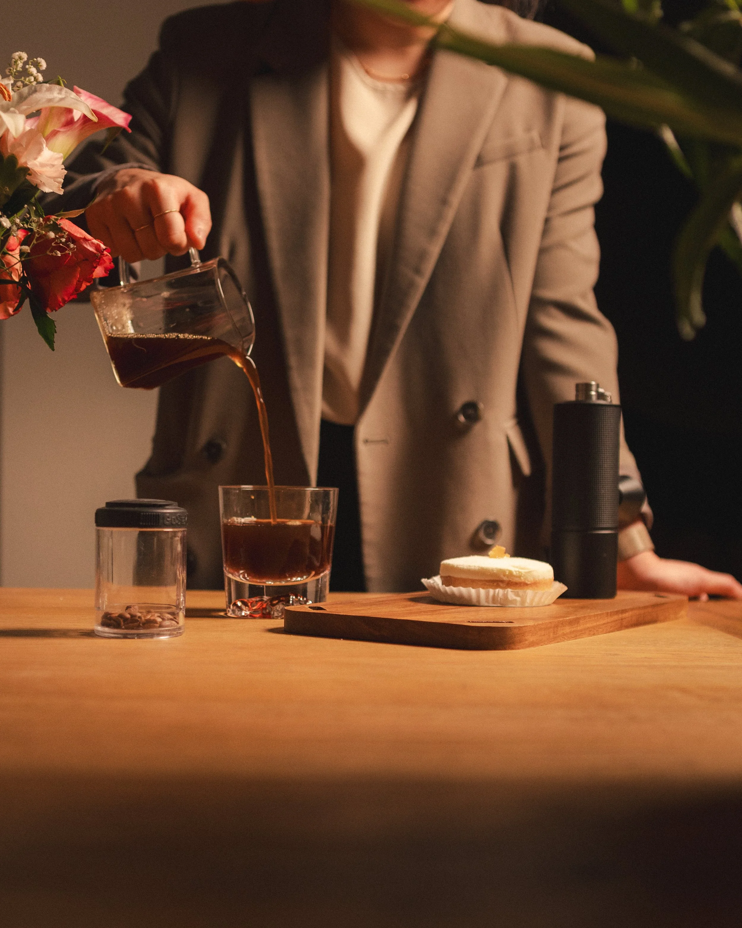 A person in a beige blazer with dark buttons pours coffee from a glass pot into a glass. The table has a wooden coaster with a cupcake on it, and a black thermos. There are flowers in the foreground.