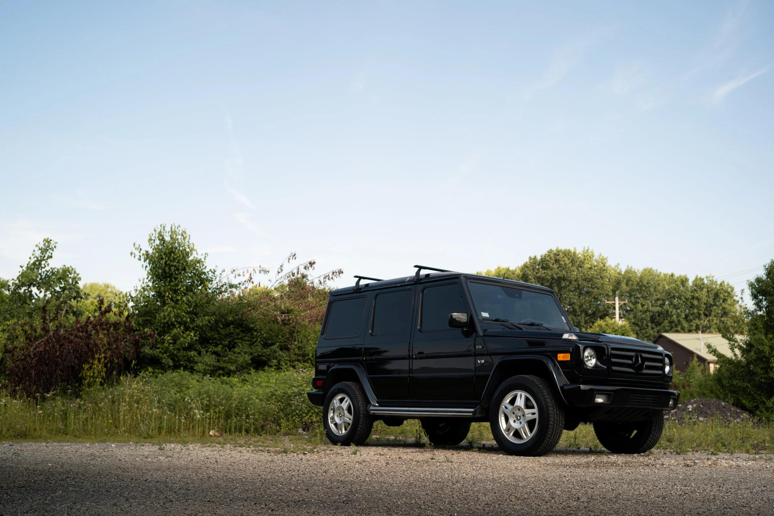 A black Mercedes-Benz G-Class SUV parked on the side of a gravel road with green trees and houses in the background under a clear blue sky.