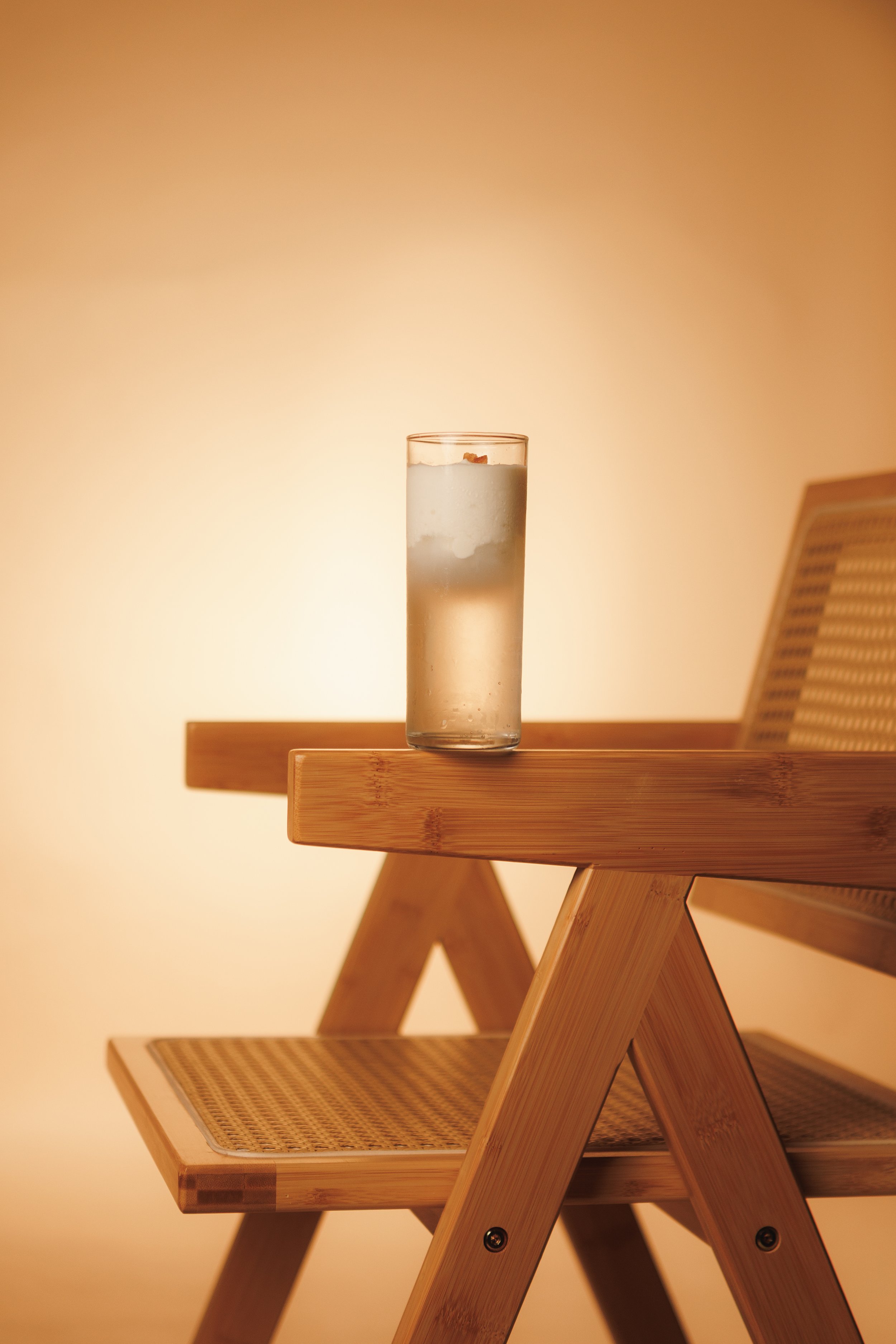 A tall glass of water with ice and a slice of lemon on a wooden table against a warm, beige background. The table has a modern, minimalist design with a smooth surface and angled legs.