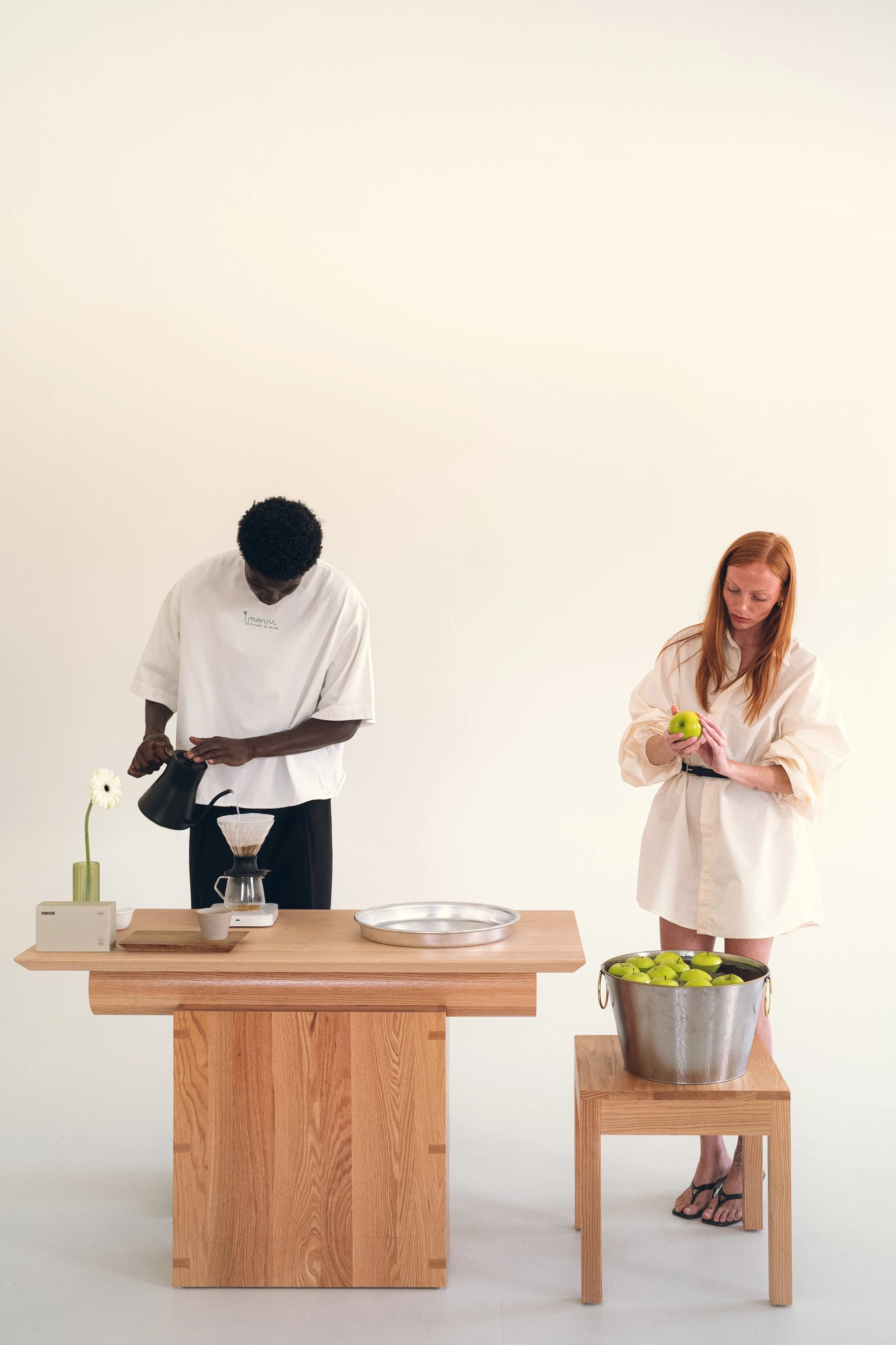 Two individuals in neutral-colored clothing standing behind wooden tables, with one pouring coffee and the other holding a green apple, against a plain light-colored background. Columbus, Ohio, David Kim Photography