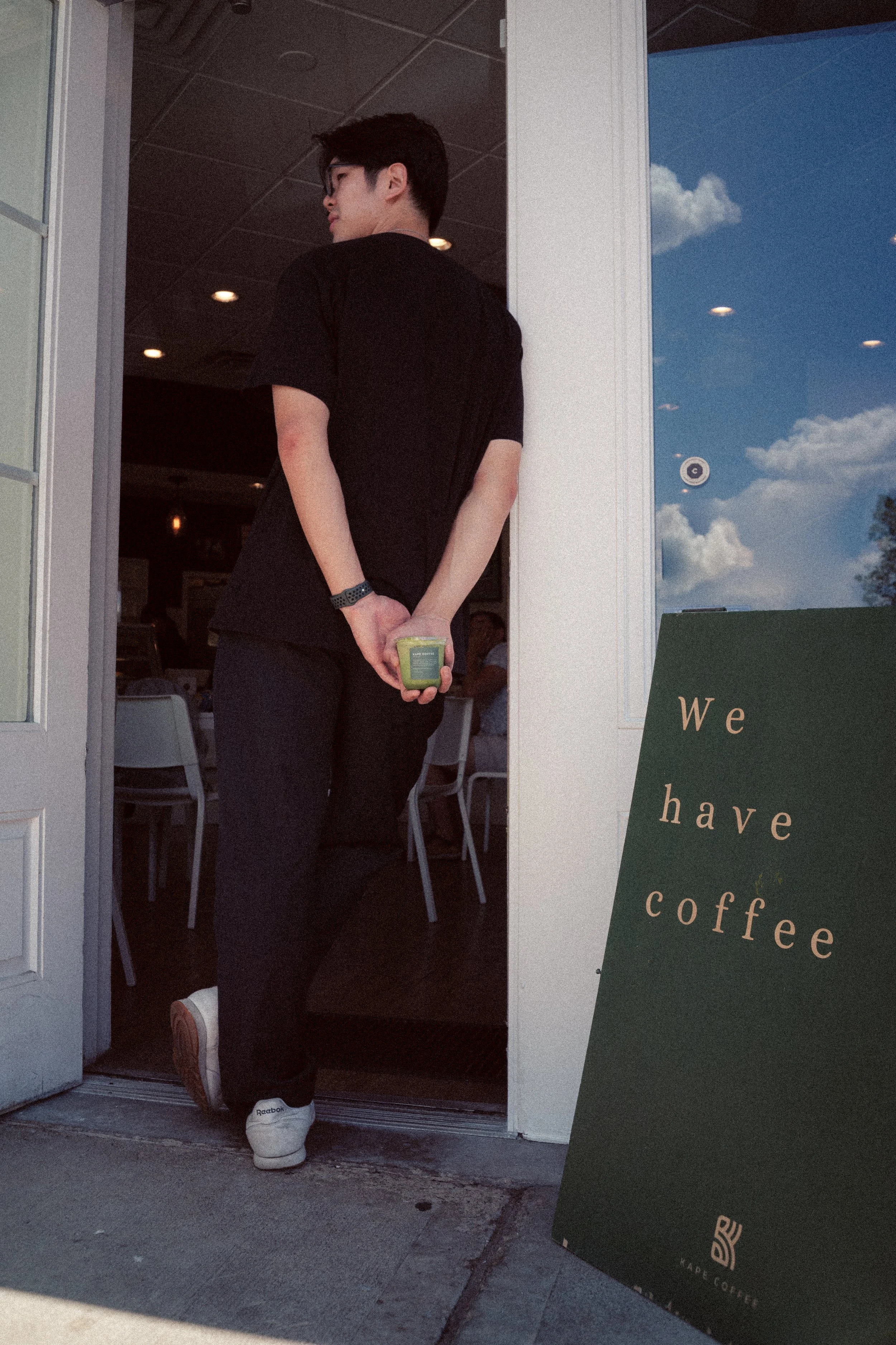 A person standing at the doorway of a coffee shop, holding a cup behind their back. The person is wearing glasses, a black t-shirt, and white sneakers. The coffee shop sign outside reads: "We have coffee." The interior has a tiled ceiling and some wa