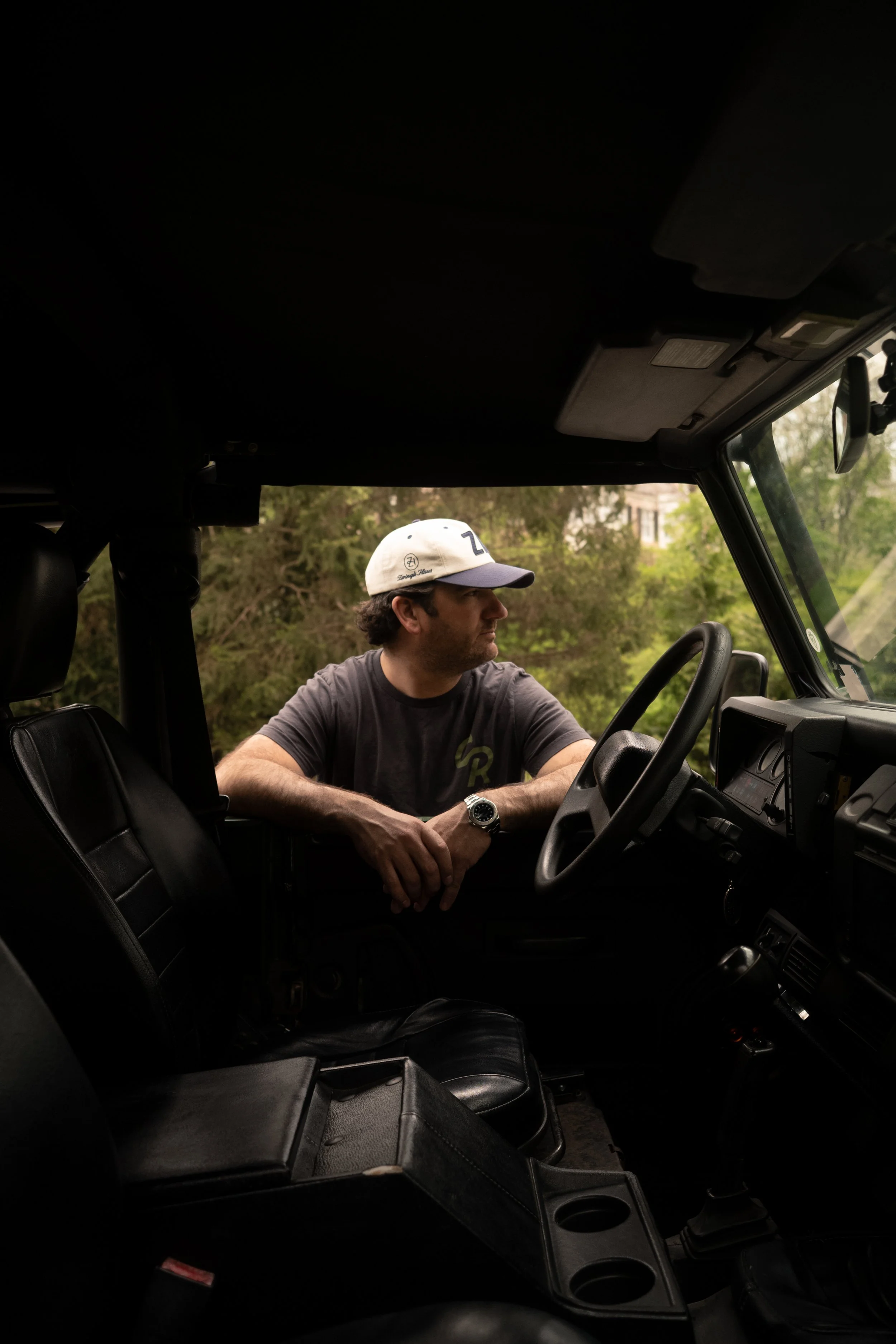A man with dark curly hair and a watch on his wrist sits in the passenger seat of a vehicle, wearing a baseball cap and a gray T-shirt. The vehicle’s interior is dark, and outside the window, green trees are visible in a suburban neighborhood.