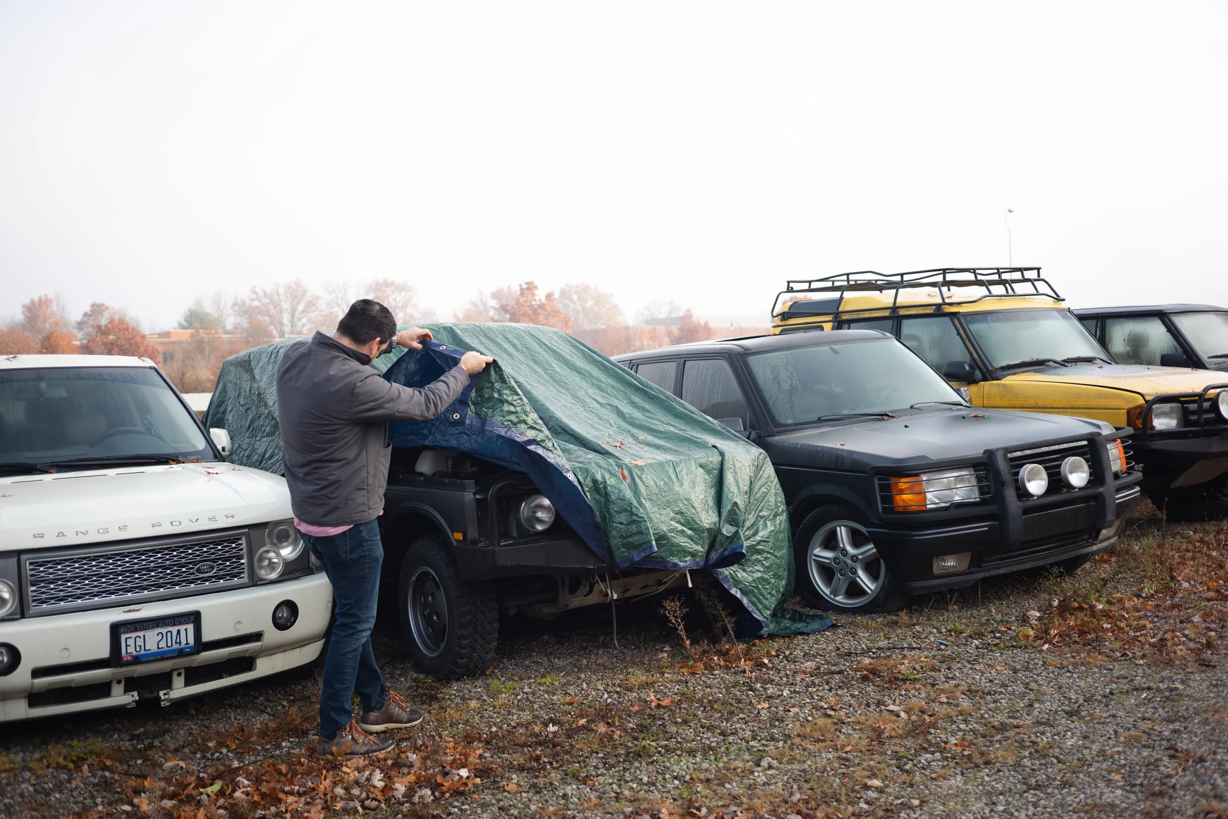 A man is removing a tarp from a black vehicle in a parking lot with several other parked cars, including white, gray, and yellow SUVs, on a cloudy day with trees in the background and fallen leaves on the ground.