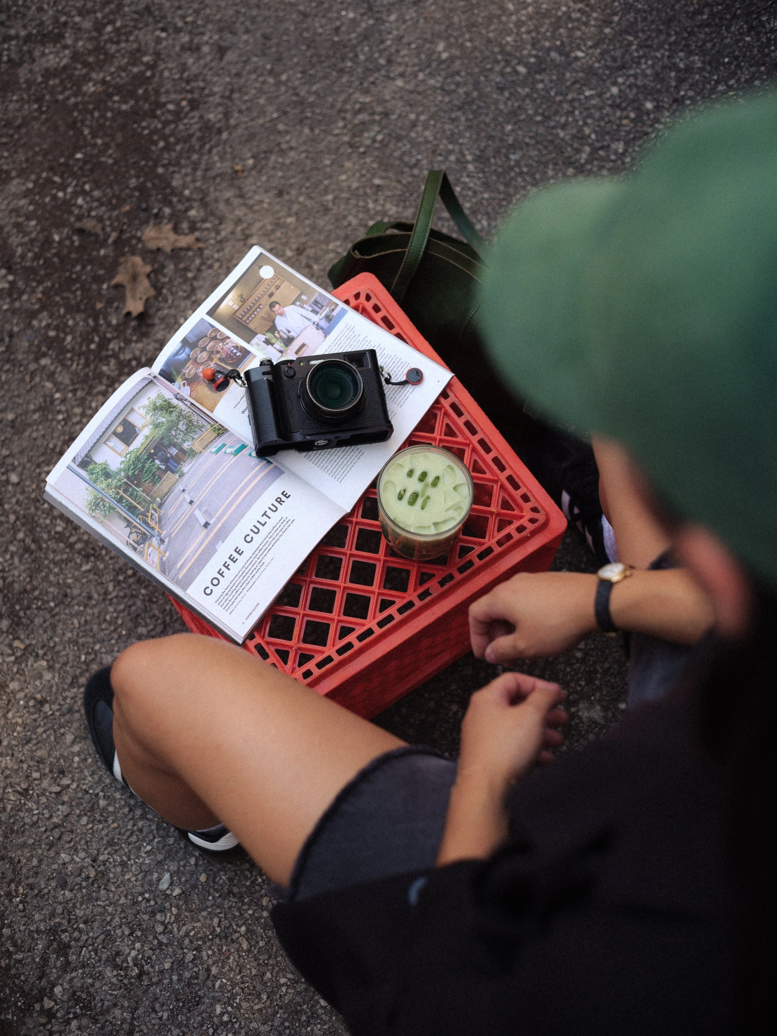 A person sitting on a red plastic crate on a gravel ground, with a magazine, a camera, and a drink nearby. The person is wearing black sneakers and has a watch on their left wrist.