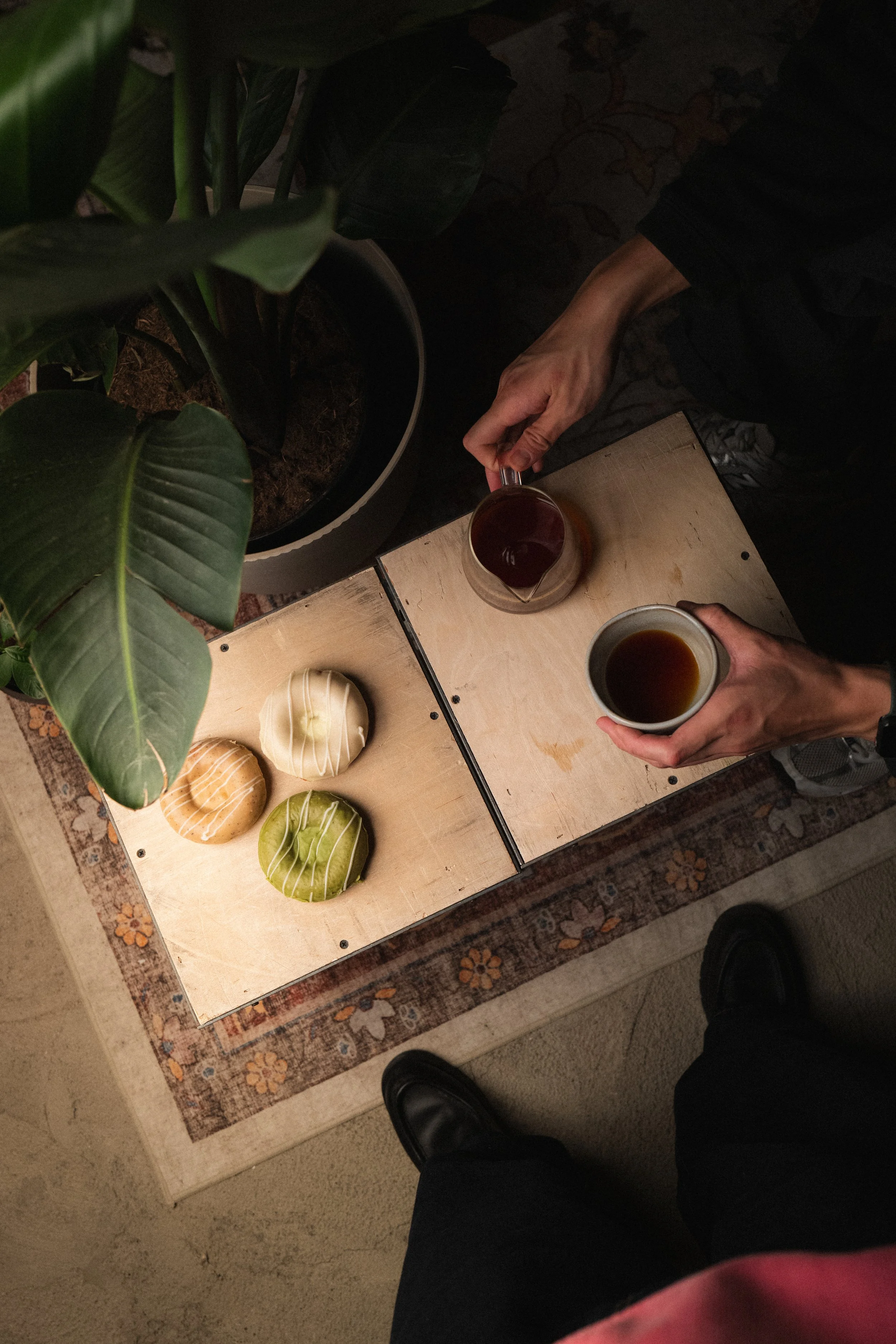 Top-down view of a person holding two coffee mugs, one in each hand, standing in front of a light wood table with four decorative balls and a potted plant nearby. The floor is underneath a patterned rug. Columbus, Ohio, David Kim Photography