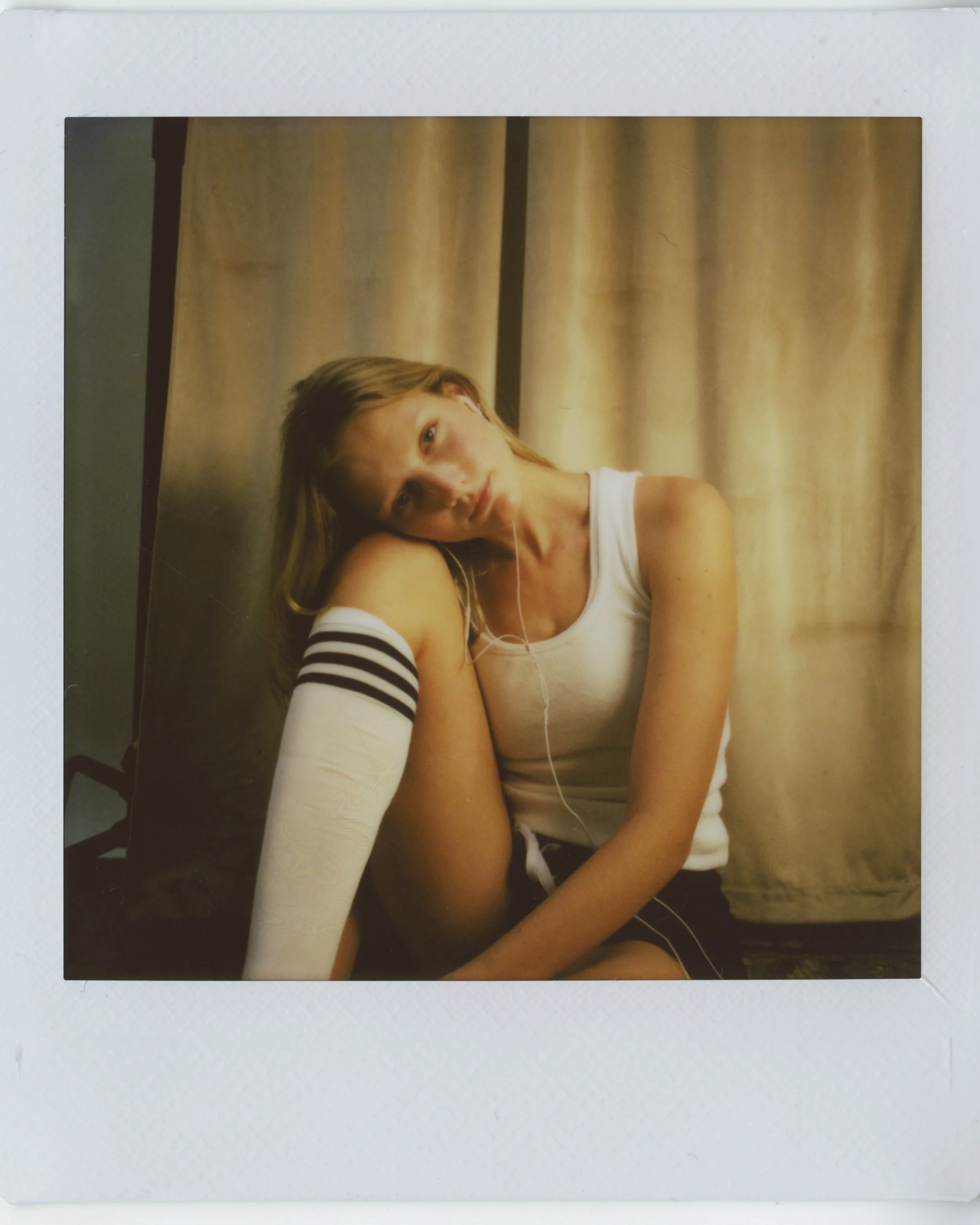 A young woman with long blonde hair, wearing a white tank top with black stripes on the sleeves, sitting in front of beige curtains, resting her head on her knee, with a neutral expression.