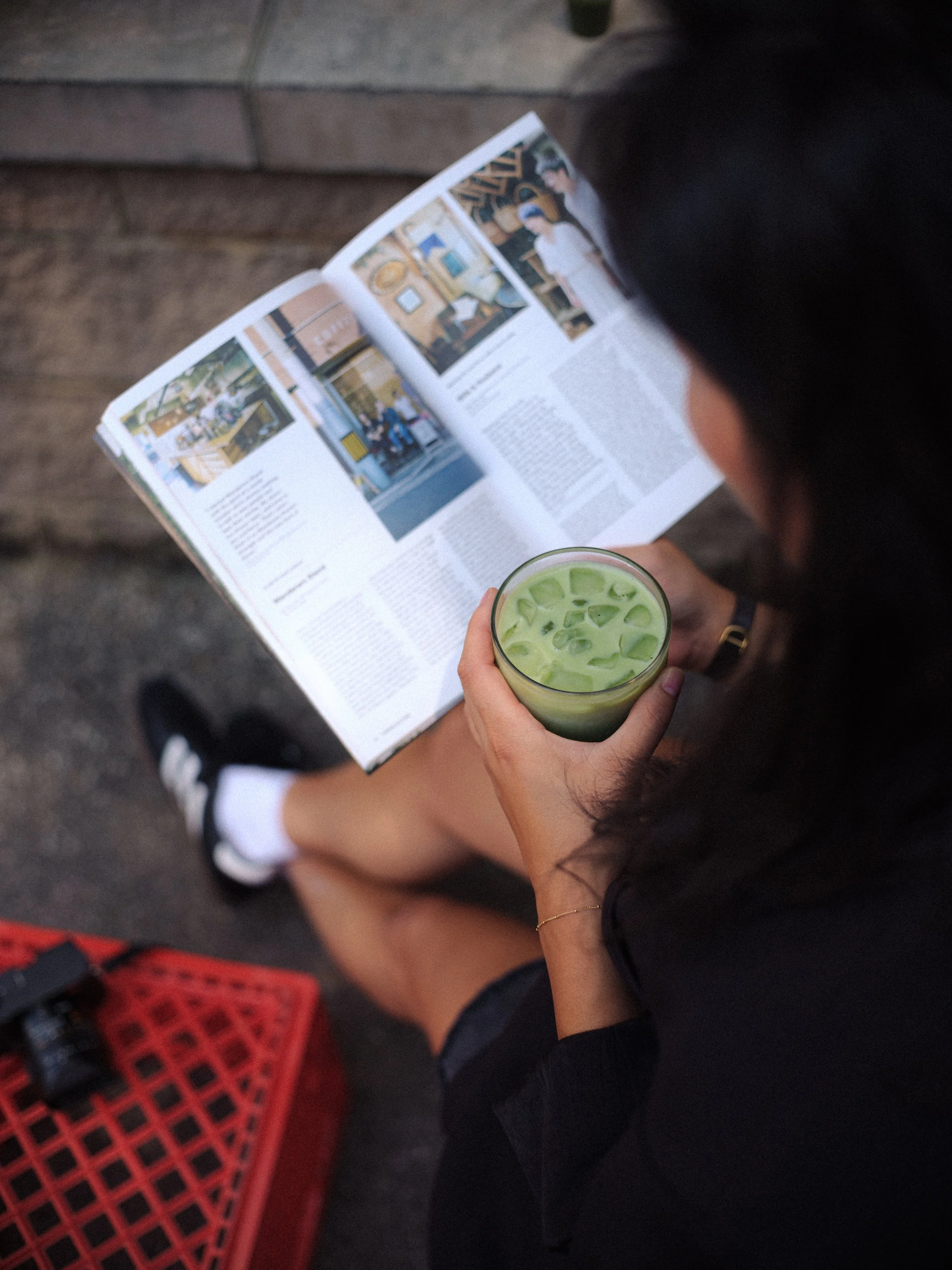 A person sitting down, holding a glass of iced green tea or matcha with ice cubes, while reading a magazine or newspaper. They are wearing black shorts and a dark top, with a black hairstyle. The background shows a concrete or stone floor and a red p