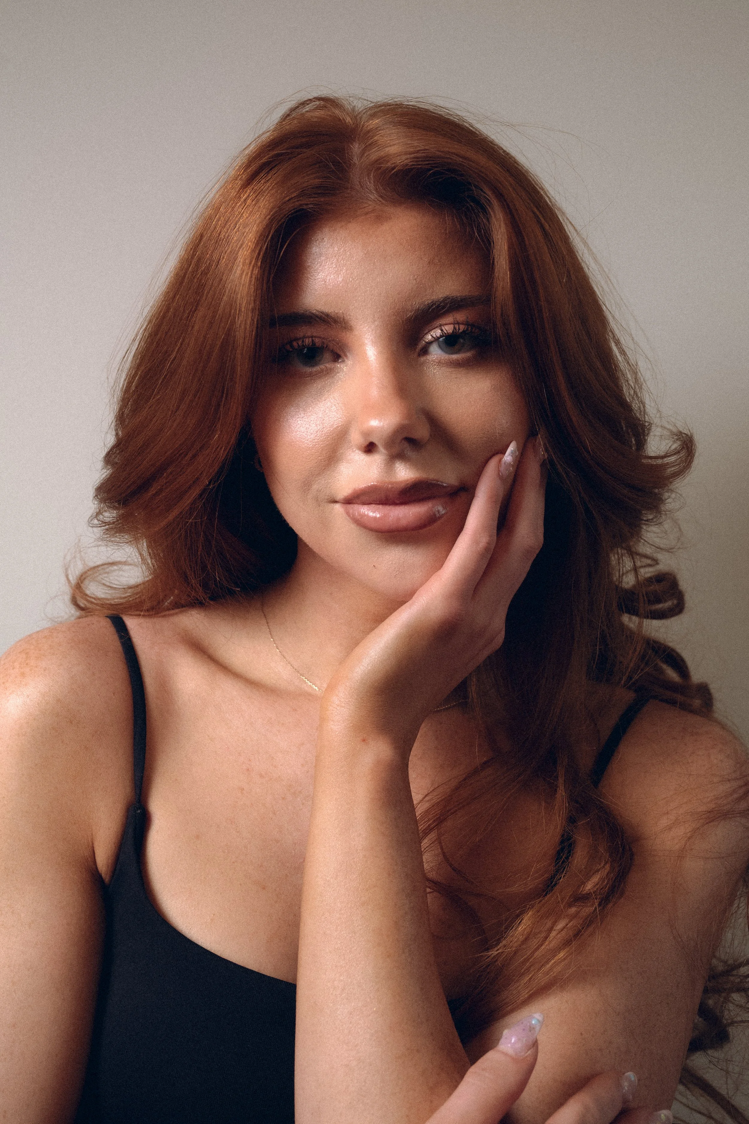 Close-up portrait of a woman with long, wavy, reddish-brown hair, wearing a black spaghetti strap top, with her hand resting on her face, and a neutral background. Columbus, Ohio, David Kim Photography