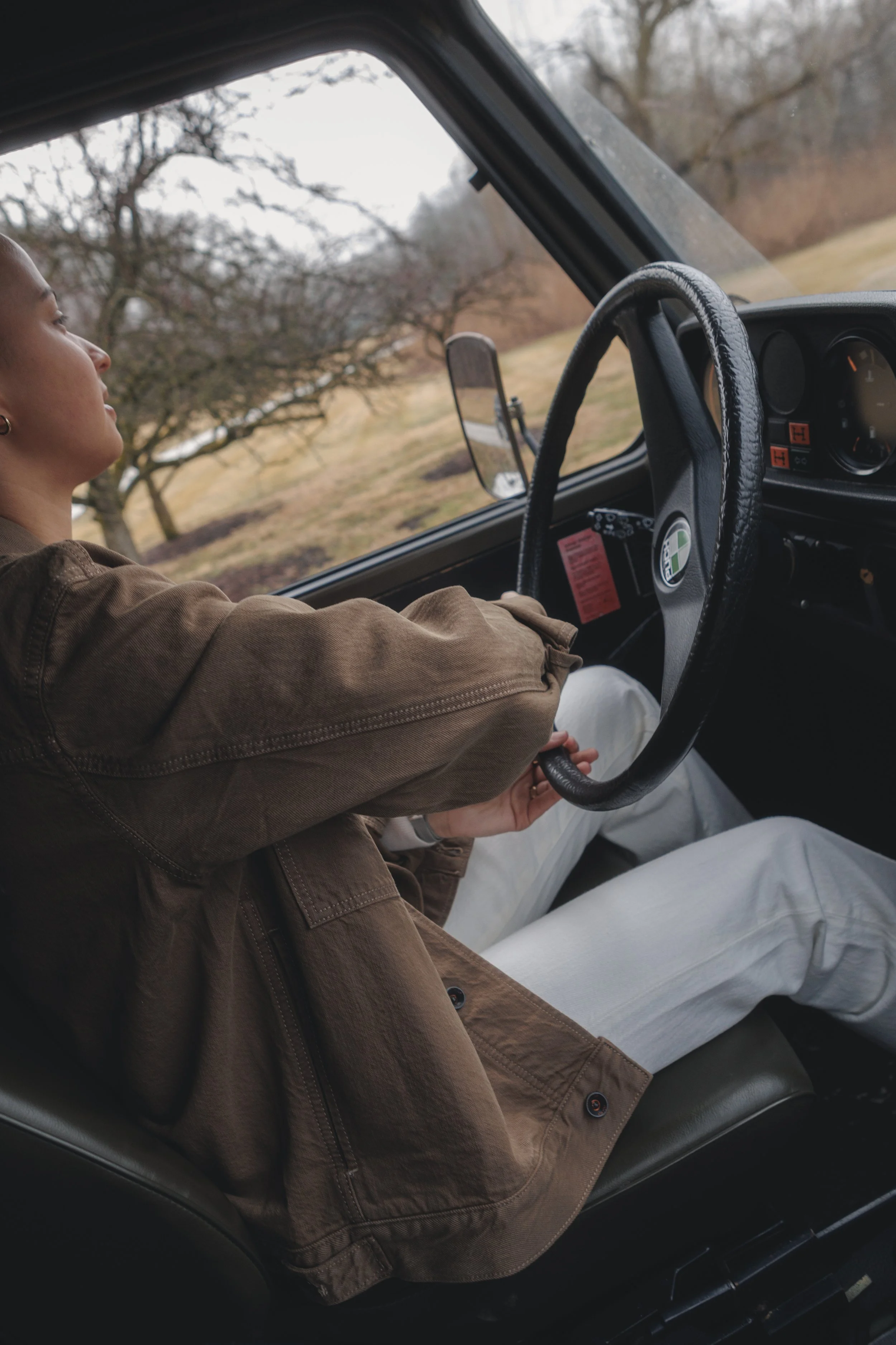 A woman is sitting in the driver seat of a vehicle during daytime, with her left arm resting on the door. The vehicle is moving through a rural area with leafless trees and grassy fields visible outside the window. The dashboard, steering wheel, and 