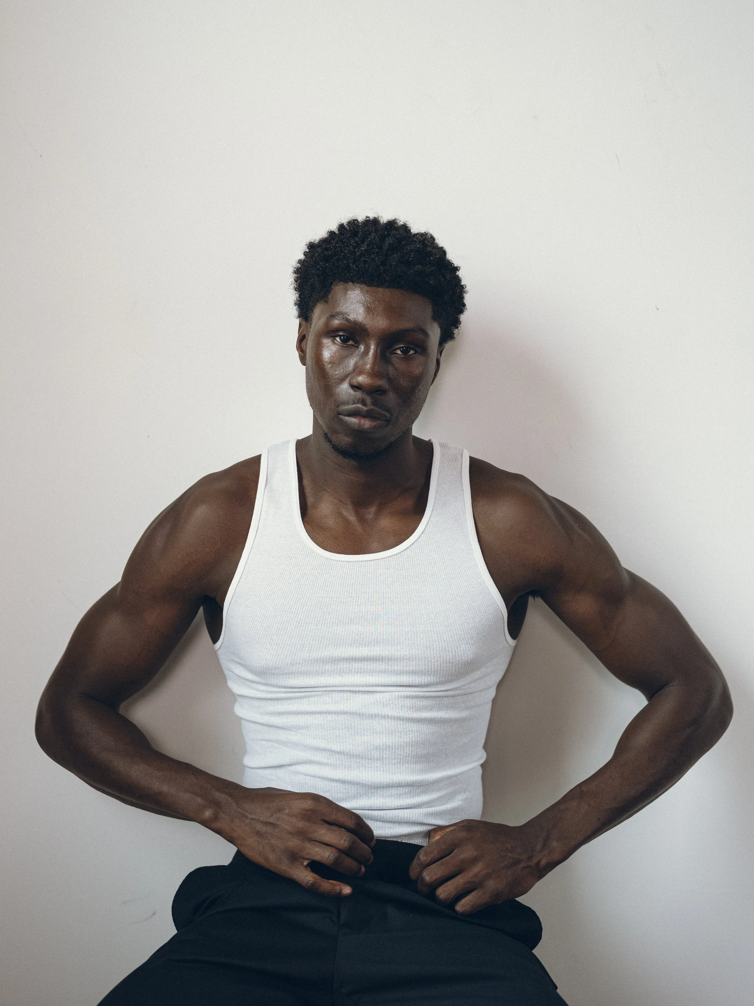 A man with dark skin, short curly hair, wearing a white tank top, sitting against a plain white wall with arms resting on his thighs.