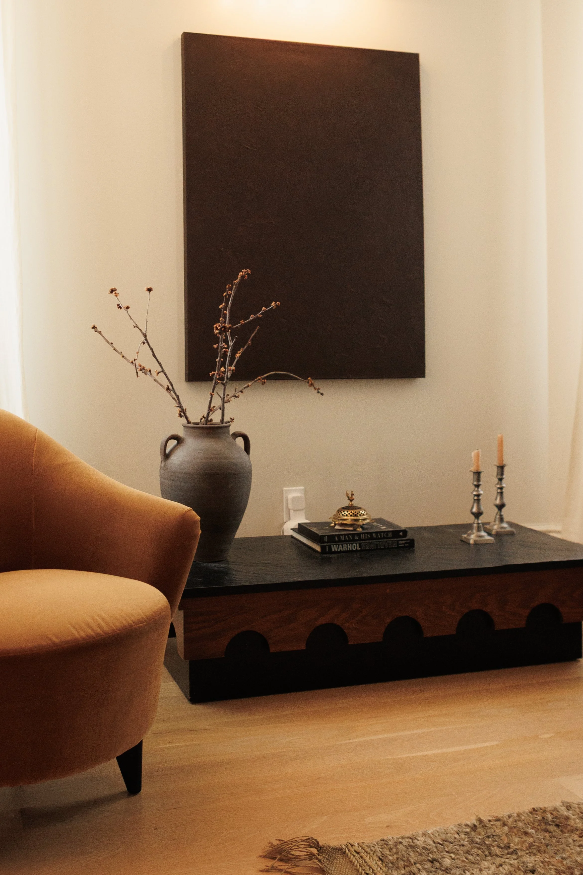 Living room with a black wooden table, a tan velvet armchair, a large grey ceramic vase with dried branches, a black tray with books and decorative items, and a black abstract wall art piece on a beige wall.