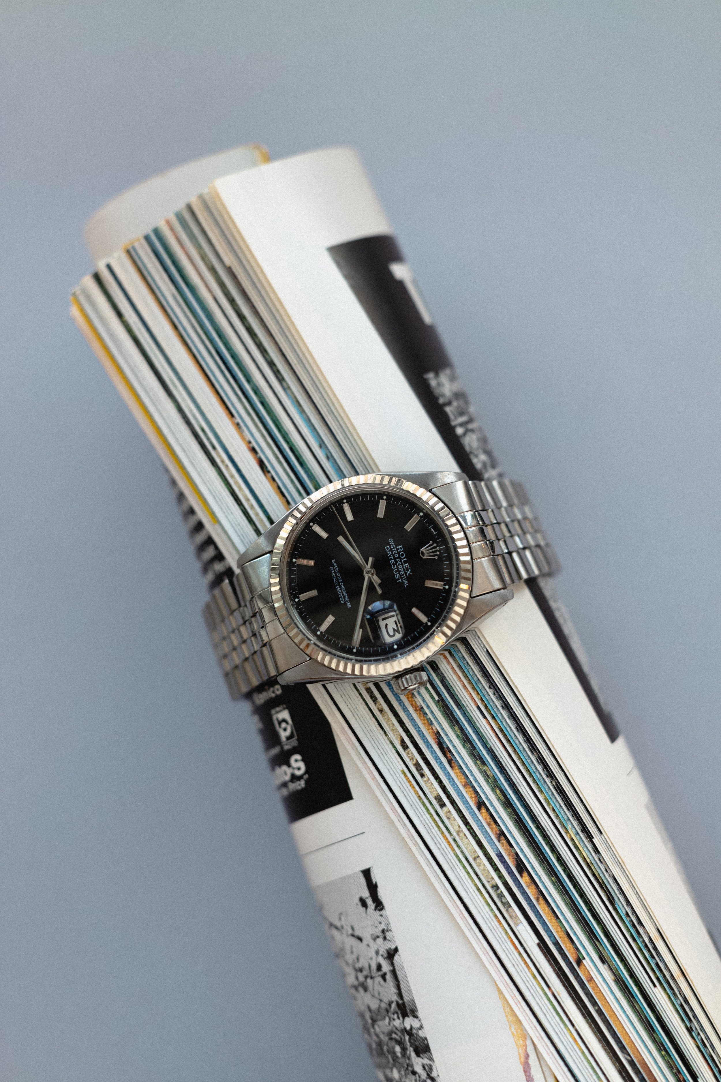 Close-up of a wristwatch with a black face and silver bracelet, resting on a rolled-up magazine or newspaper.