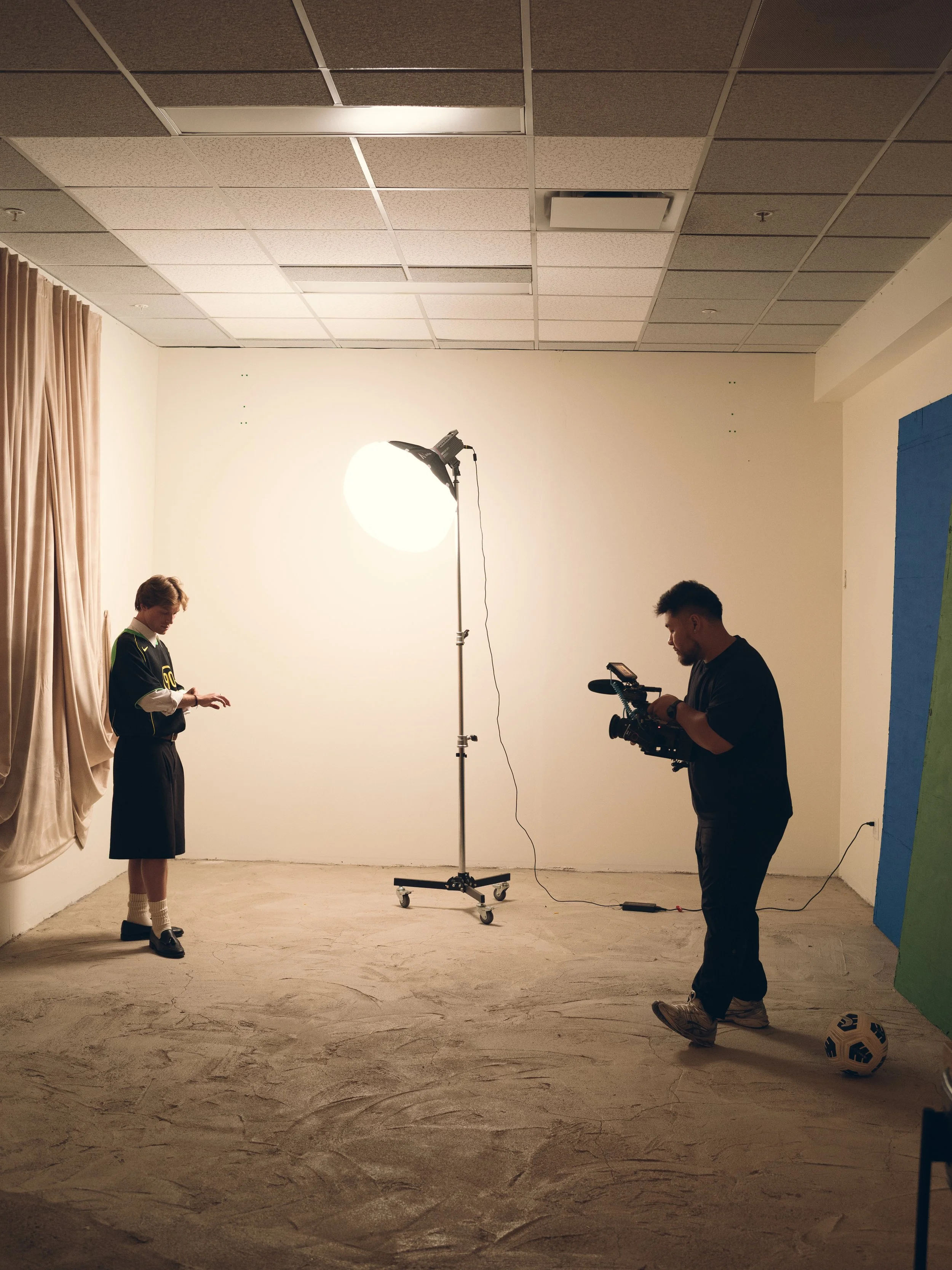 Behind the scenes of a video shoot with two men. One in a black T-shirt with a camera and another in a sporty dress with sneakers. A large studio light is set up, and a soccer ball on the ground. Columbus, Ohio, David Kim Photography