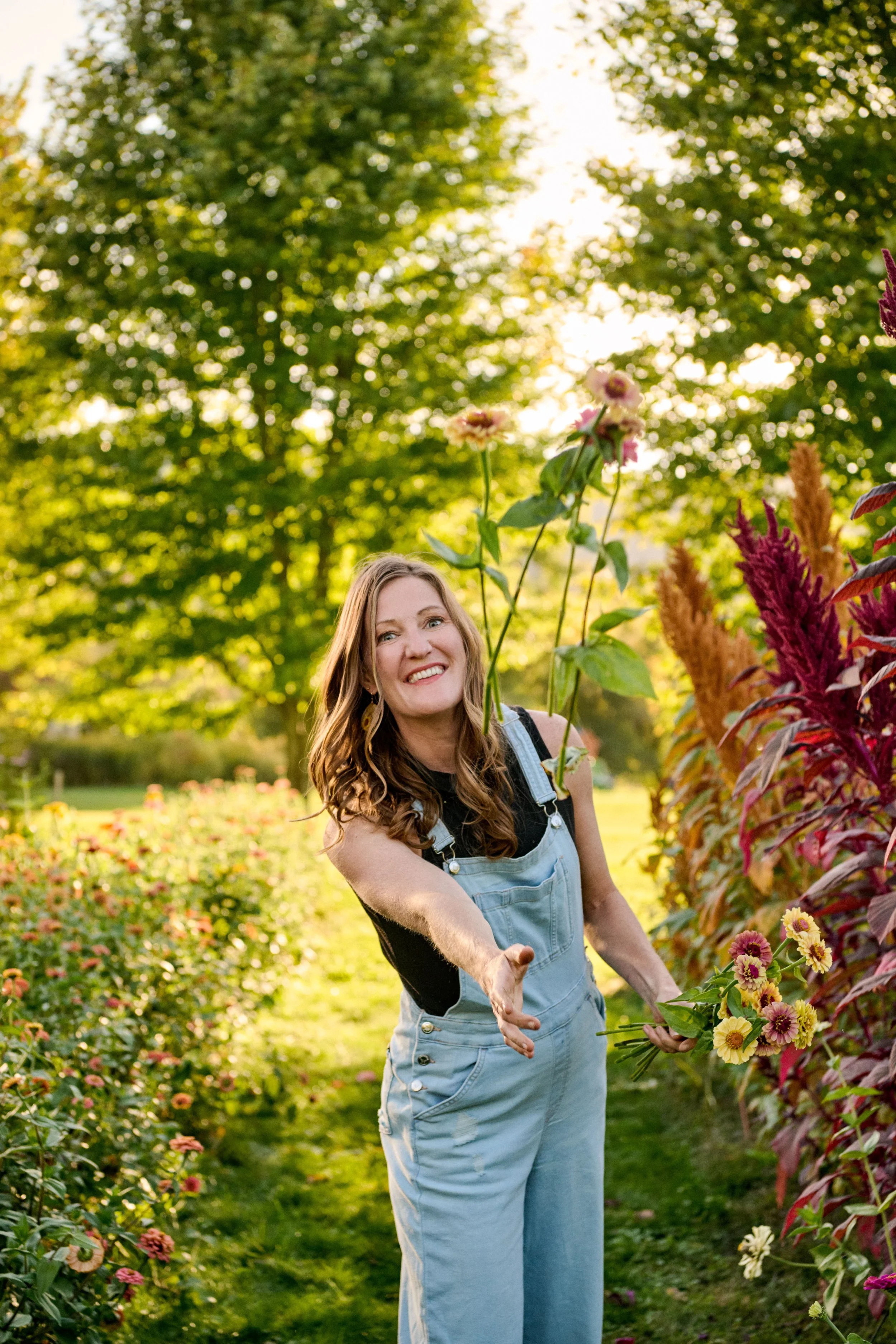A woman (Laura) with long wavy hair smiling and reaching out her hand in a garden with colorful flowers and green trees in the background during sunset.