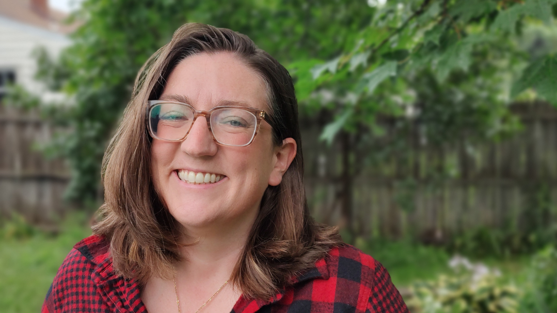 A woman with glasses smiling outdoors in a garden with green trees and a wooden fence in the background.