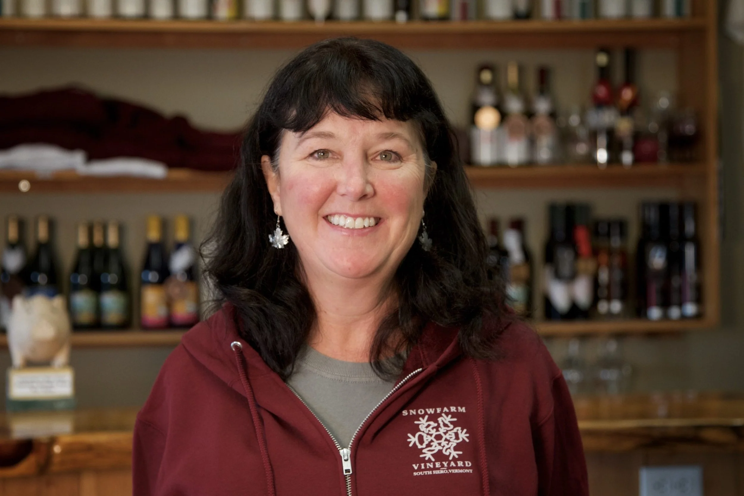 A woman with dark hair, wearing a maroon jacket with a snowflake logo, standing in a wine shop or vineyard with shelves of wine bottles in the background.