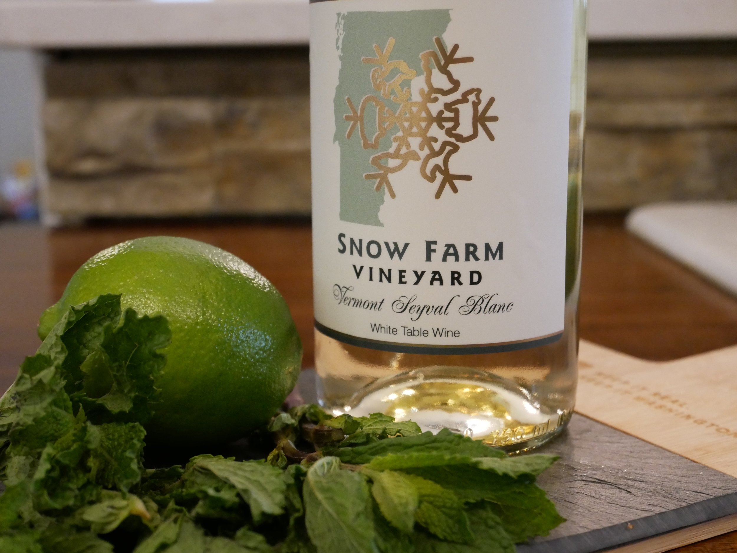 A green lime, fresh mint leaves, and a bottle of Snow Farm Vineyard Vermont Seyval Blanc white wine on a wooden surface.