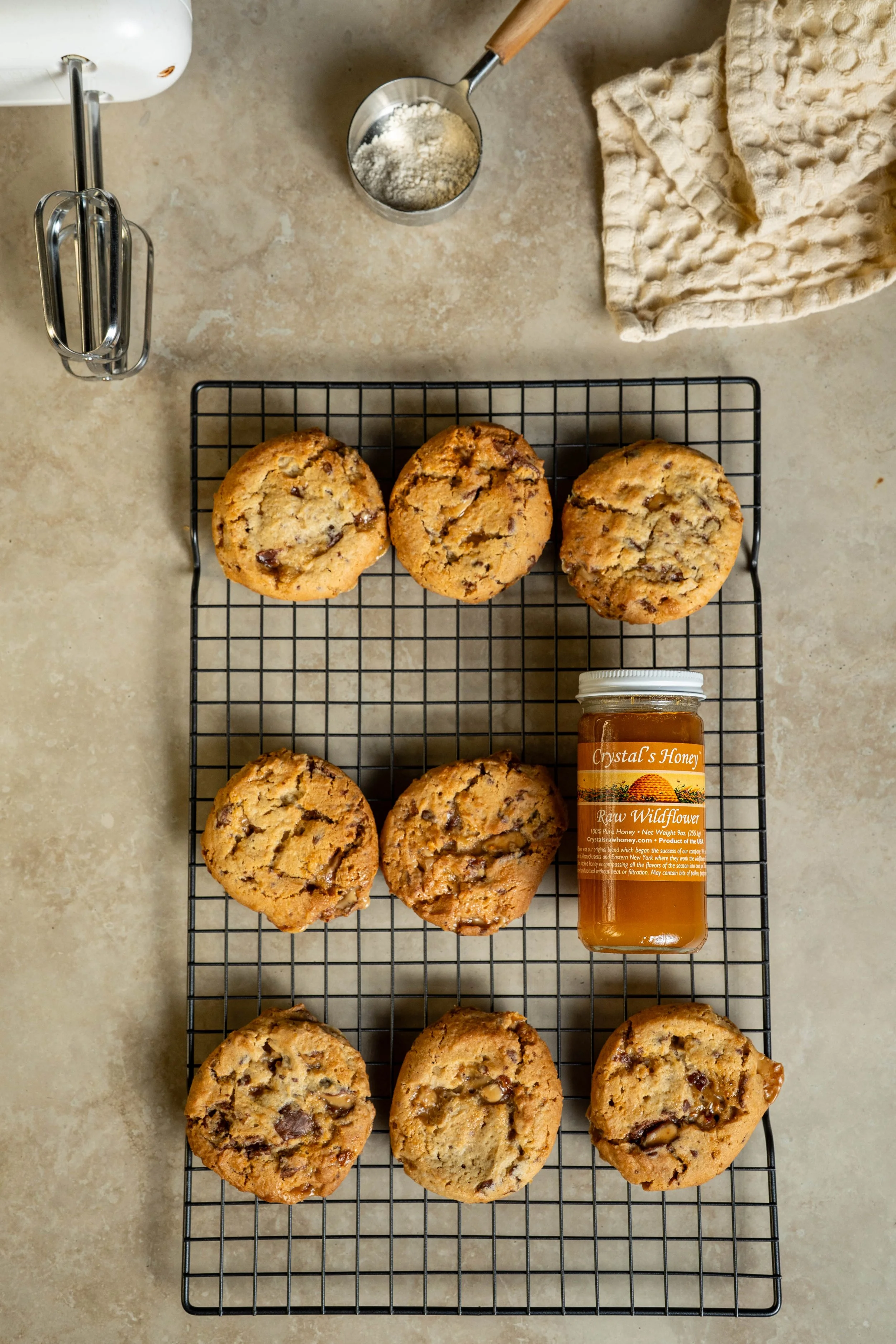 Freshly baked chocolate chip cookies on a cooling rack, a jar of Crystals Honey, and baking tools on a kitchen countertop.