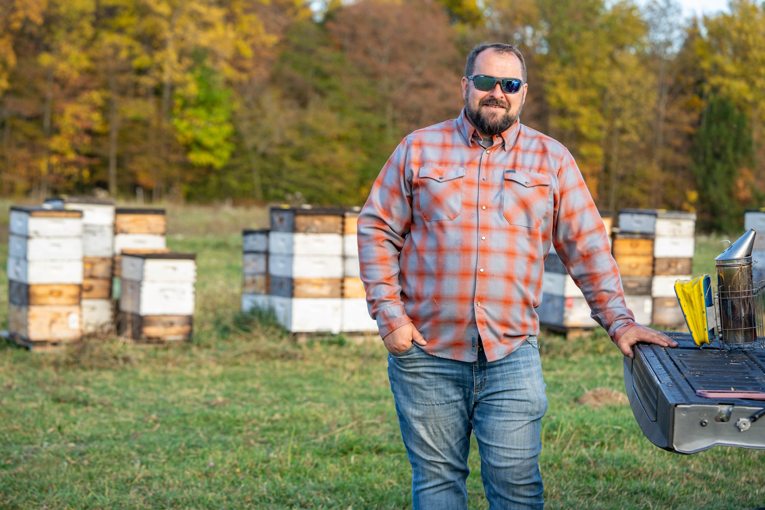 Glenn Card with sunglasses and a beard wearing a plaid shirt stands outdoors near beehives, with trees in autumn foliage in the background.