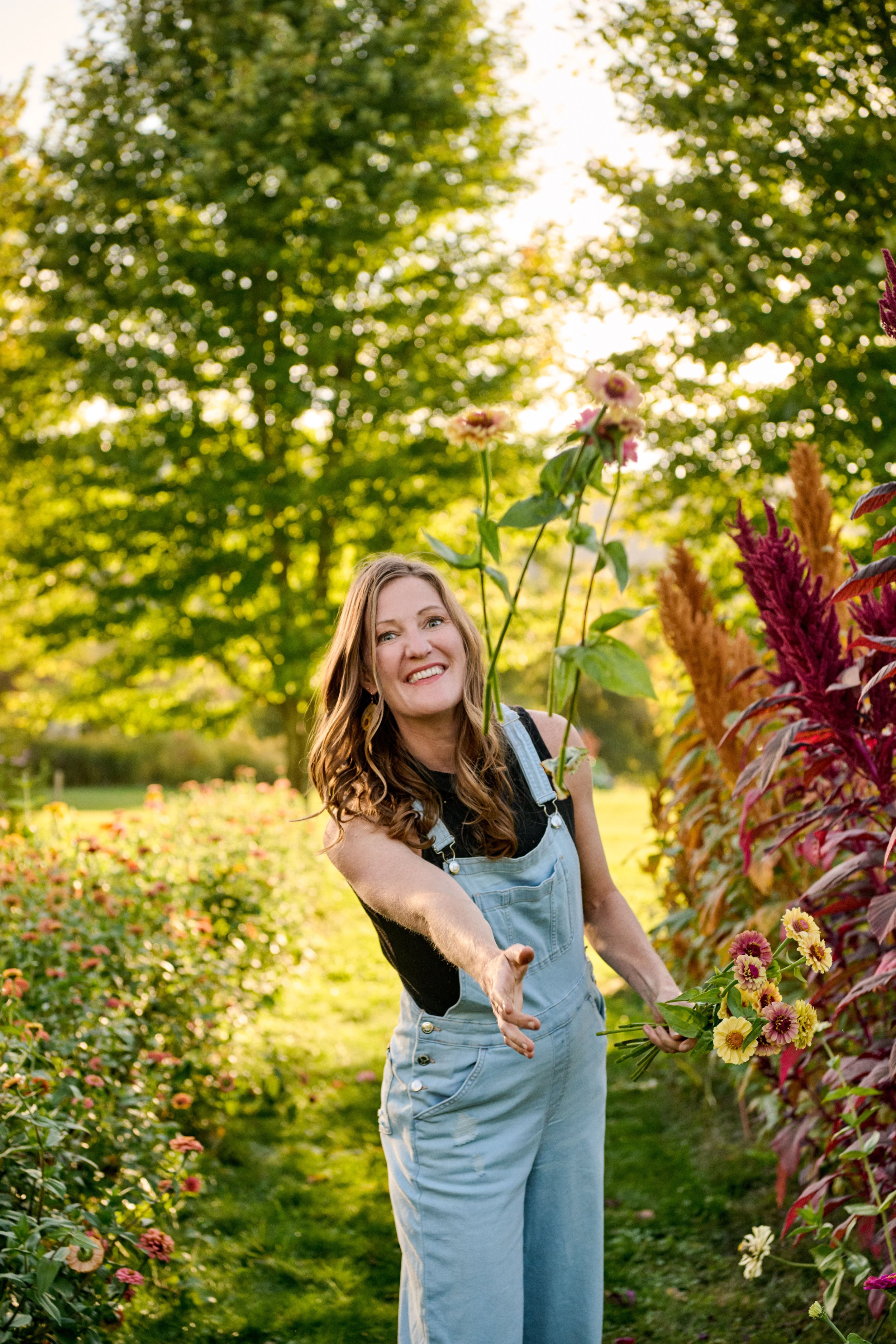A smiling woman (Laura)  in denim overalls extending her hand towards the camera in a colorful garden with flowers and trees in the background.