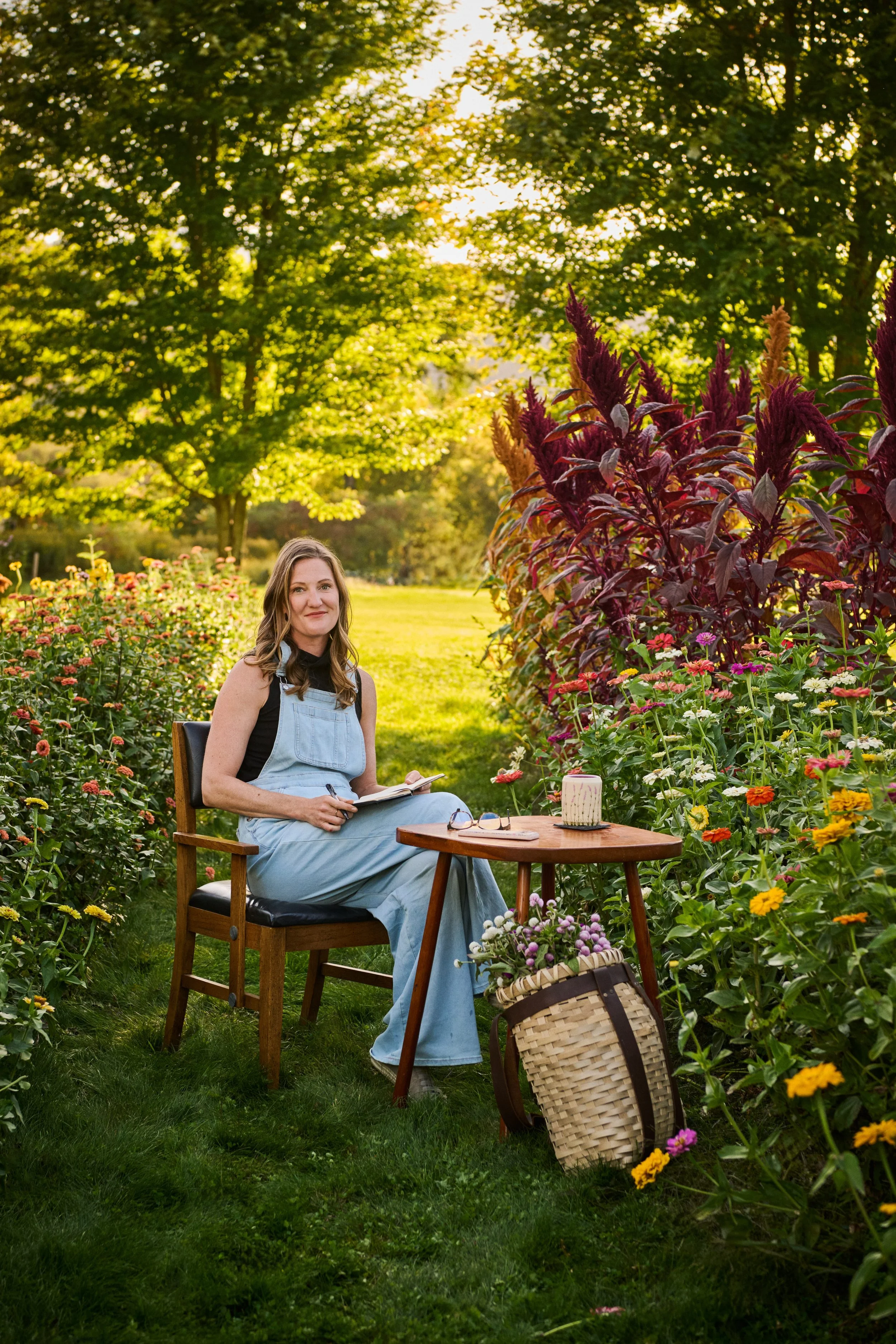 A woman (Laura) sitting on a wooden chair in a garden surrounded by colorful flowers, holding a notebook, with a basket of flowers on the ground and a small table with candles nearby, during sunset.