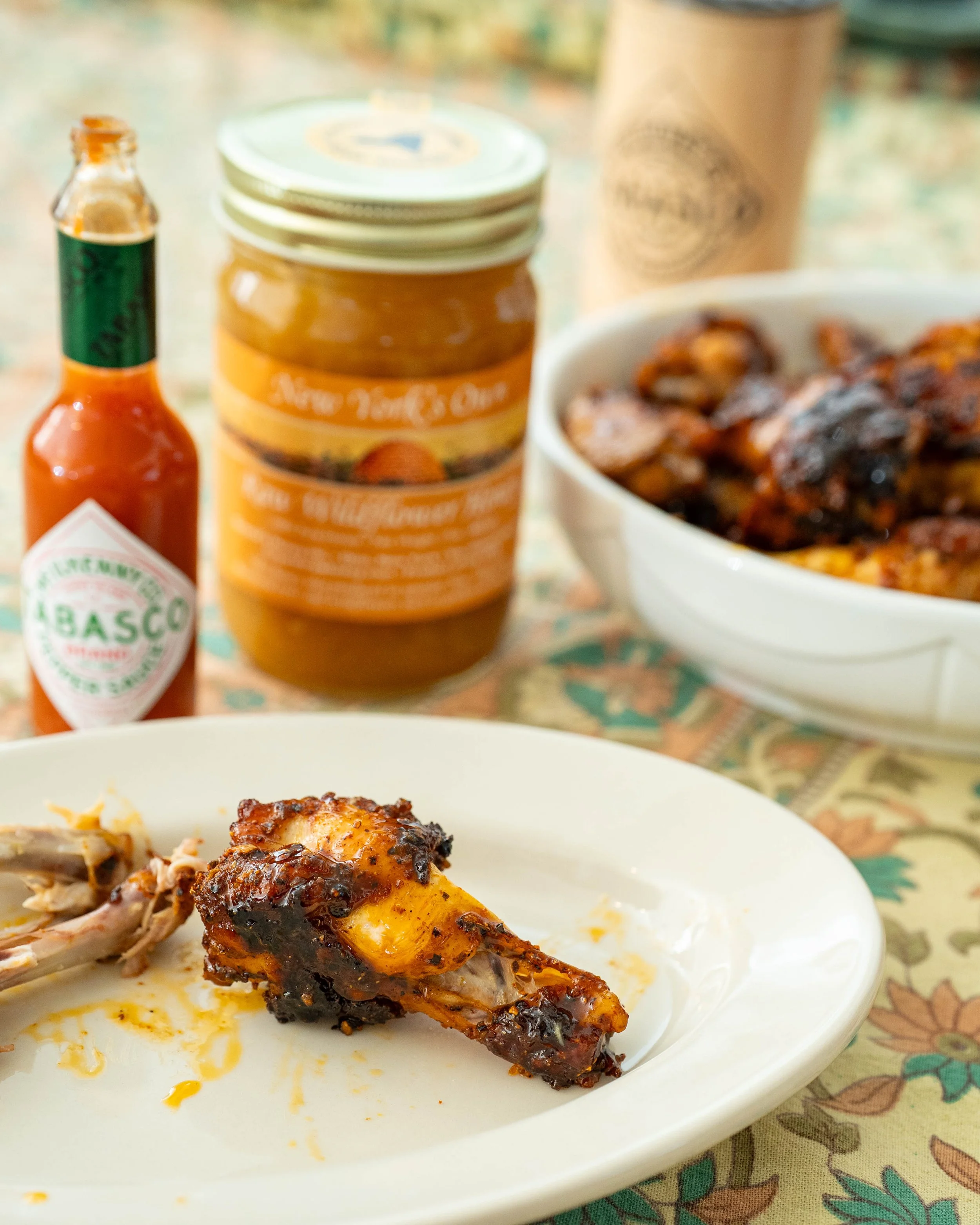 Close-up of a roasted chicken leg with crispy, glazed skin on a white plate, with some shredded chicken and melted cheese, placed on a patterned tablecloth.