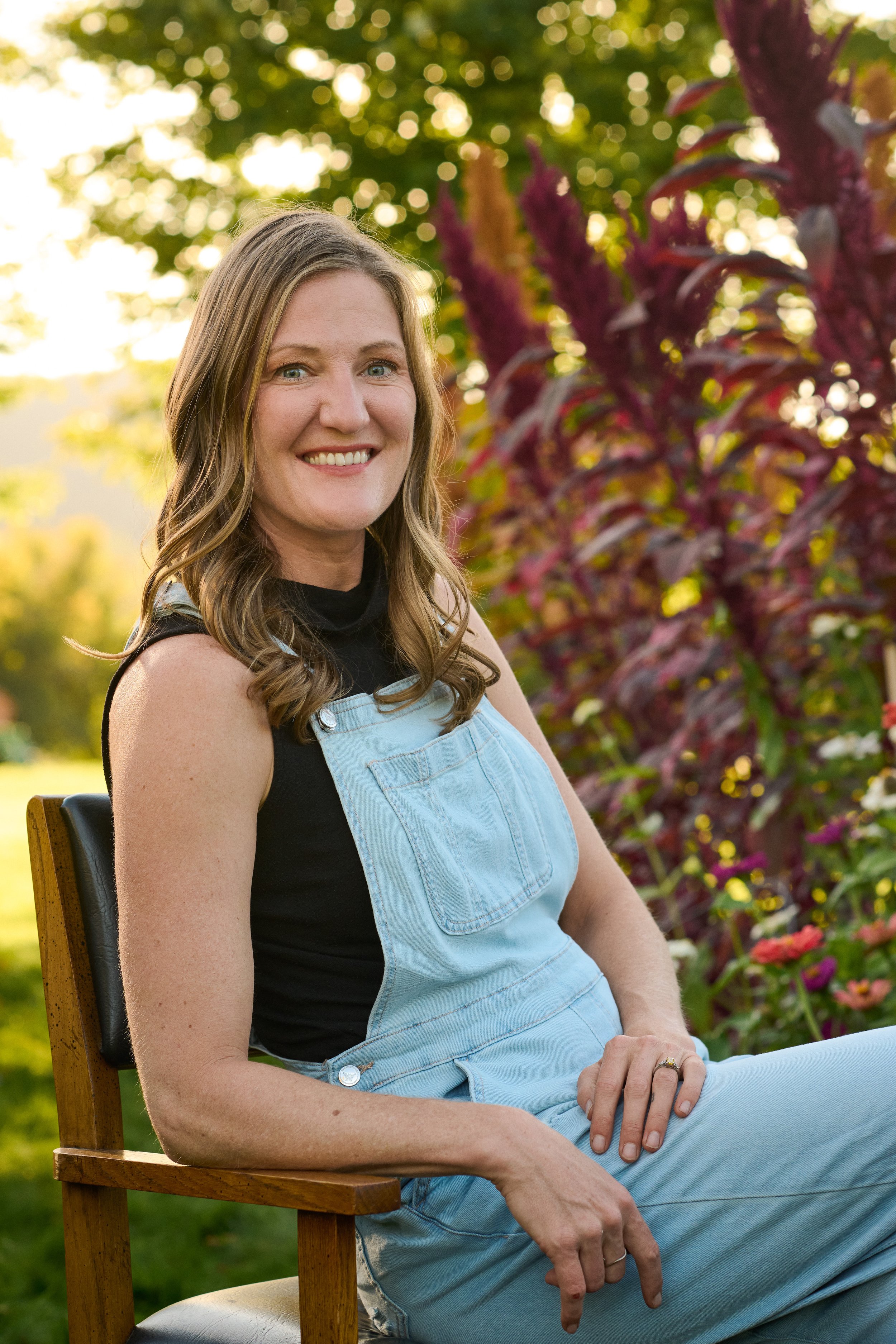 A woman with long wavy brown hair, smiling and sitting outdoors on a wooden chair, wearing a black shirt and light blue denim overalls, with colorful flowers and green foliage in the background.